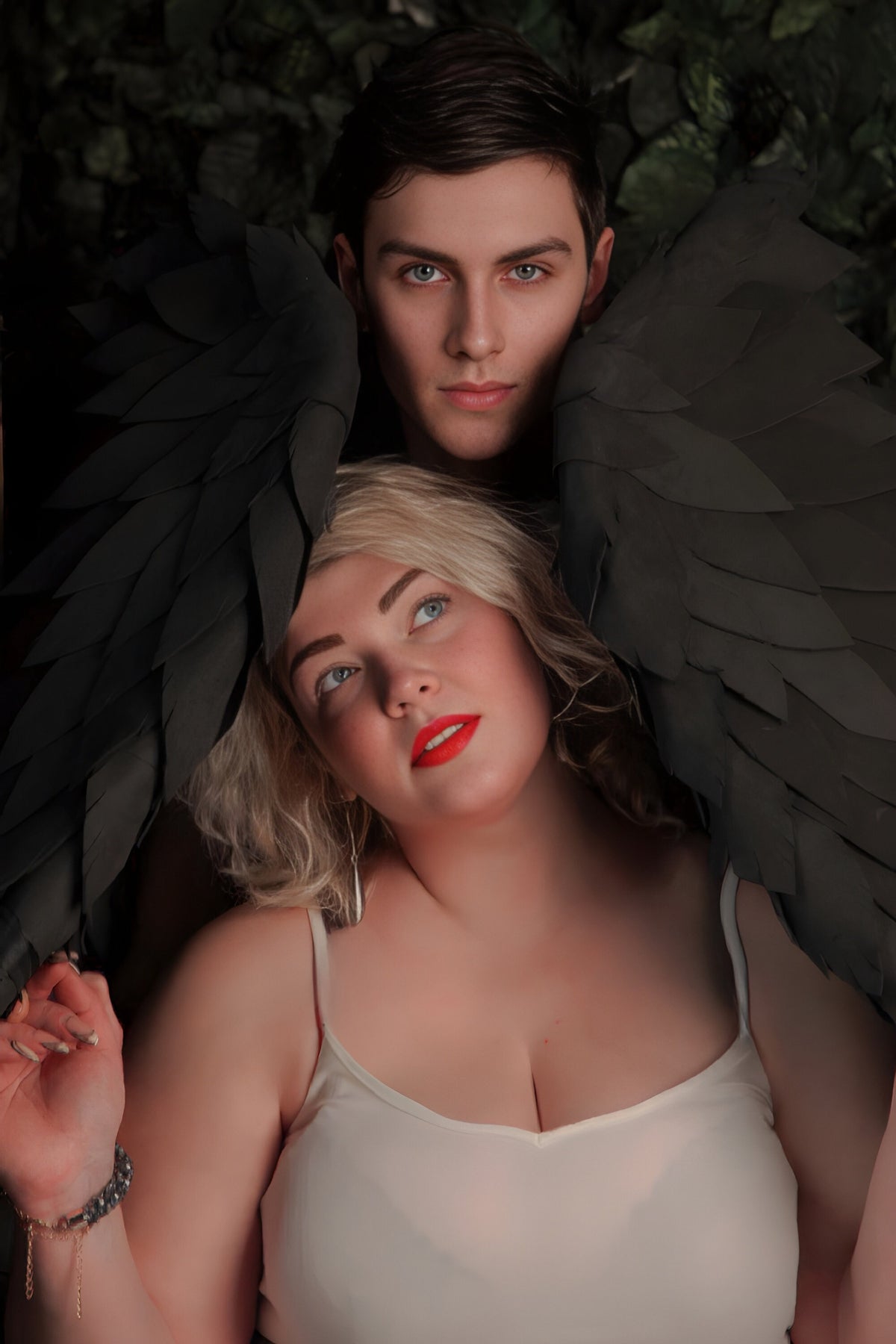 May include: A woman with blonde hair and red lipstick looks up while wearing a white tank top. A man with dark hair and a serious expression is behind her. They are both wearing black feathered wings.
