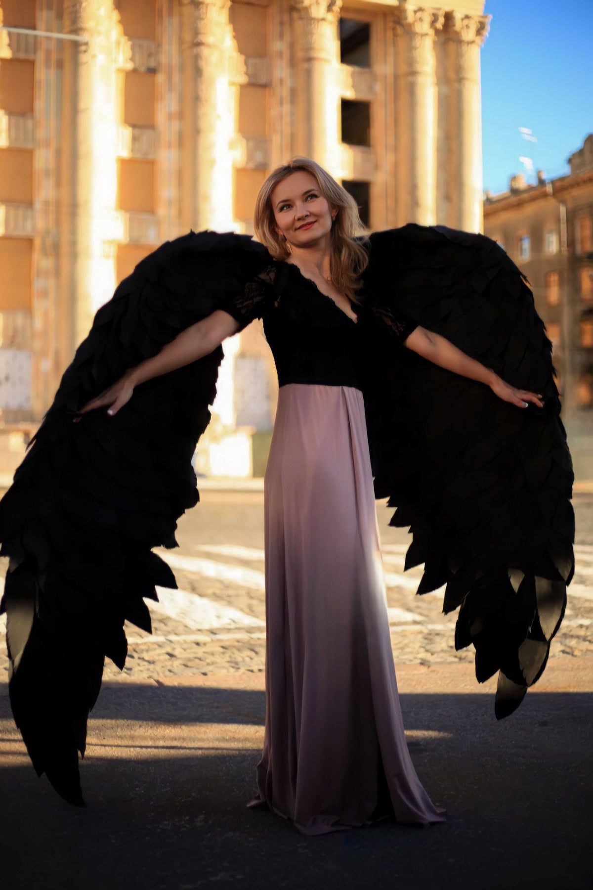 May include: A woman wearing a black lace top and a long, flowing pink skirt stands with her arms outstretched, holding large black wings. The wings are made of fabric and have a feathery texture.