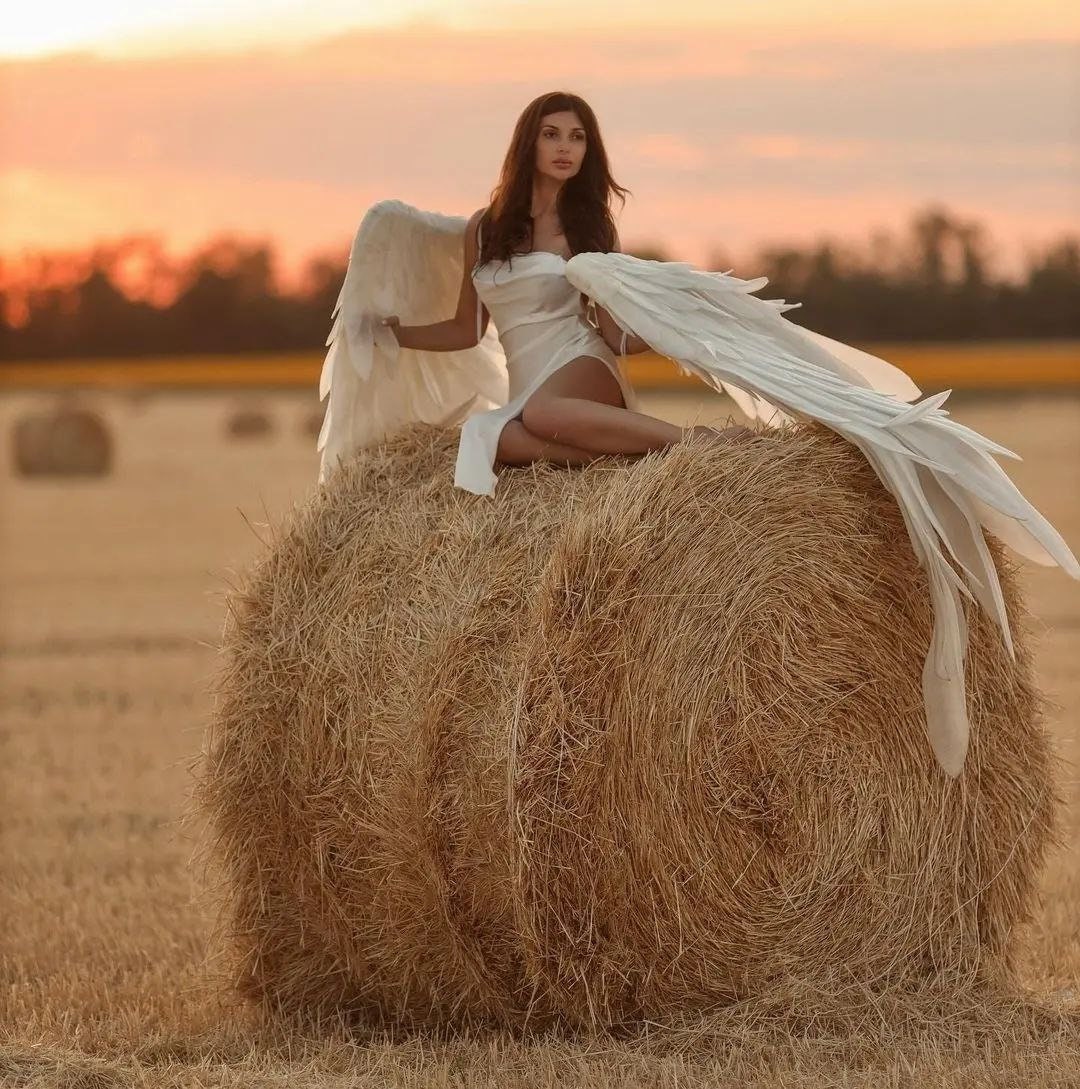 May include: A woman wearing a white dress sits on a large round bale of hay. She is holding a large white feathered angel wing. The background is a sunset.