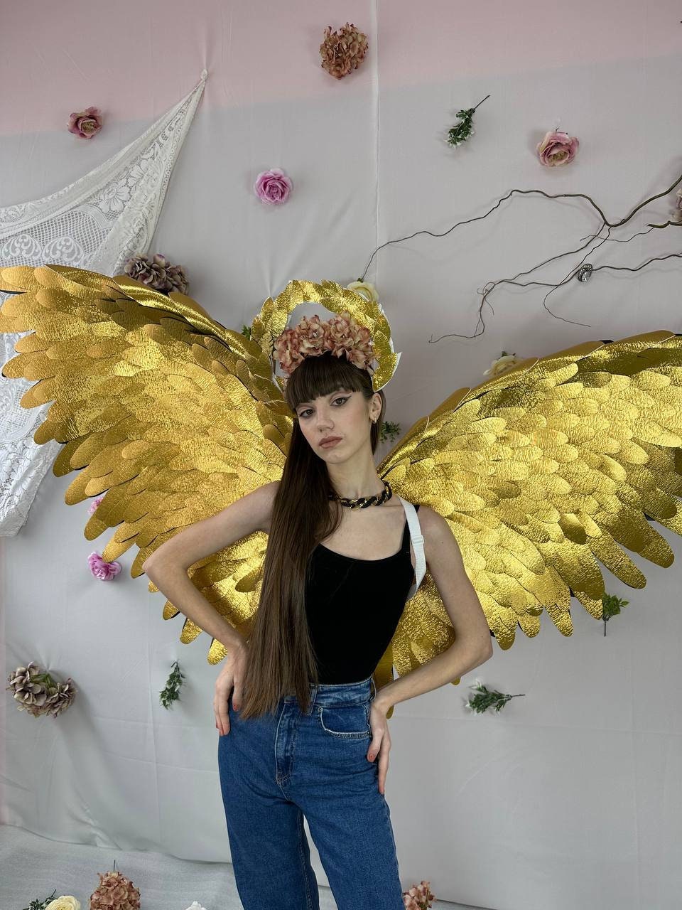 May include: A person wearing a black tank top, blue jeans, and a gold halo with flowers. They are wearing large gold angel wings.