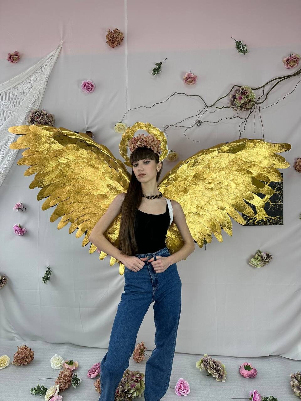 May include: A person wearing a black tank top and blue jeans with large gold angel wings and a floral halo.