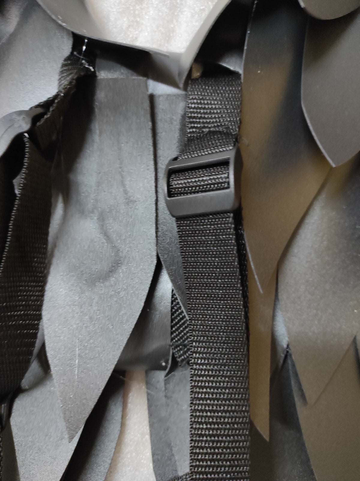 May include: Black adjustable strap with a buckle, attached to a black fabric garment. The fabric has a layered, scaly texture.