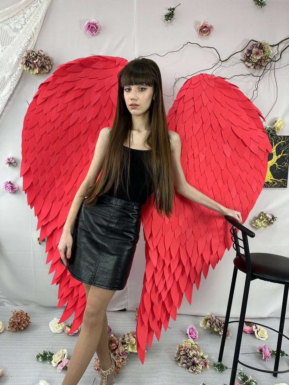 May include: A person wearing a black top and a black leather skirt stands next to large, red, feathered angel wings. The wings are the focal point, with a textured, layered design. The background includes floral decorations and a black chair.