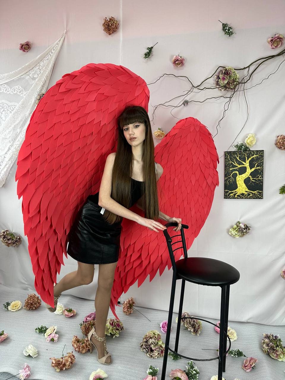 May include: A woman wearing a black skirt and heels poses with large red angel wings. The wings are made of foam and have a feathery texture.