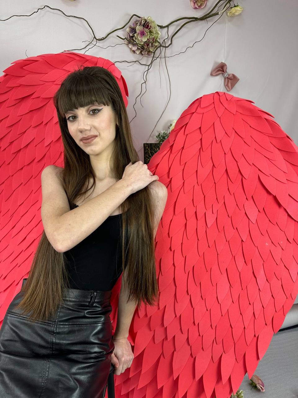 May include: A woman wearing a black top and a black leather skirt poses in front of large, vibrant red angel wings. The wings are made of layered, petal-shaped pieces. The background includes floral decorations.