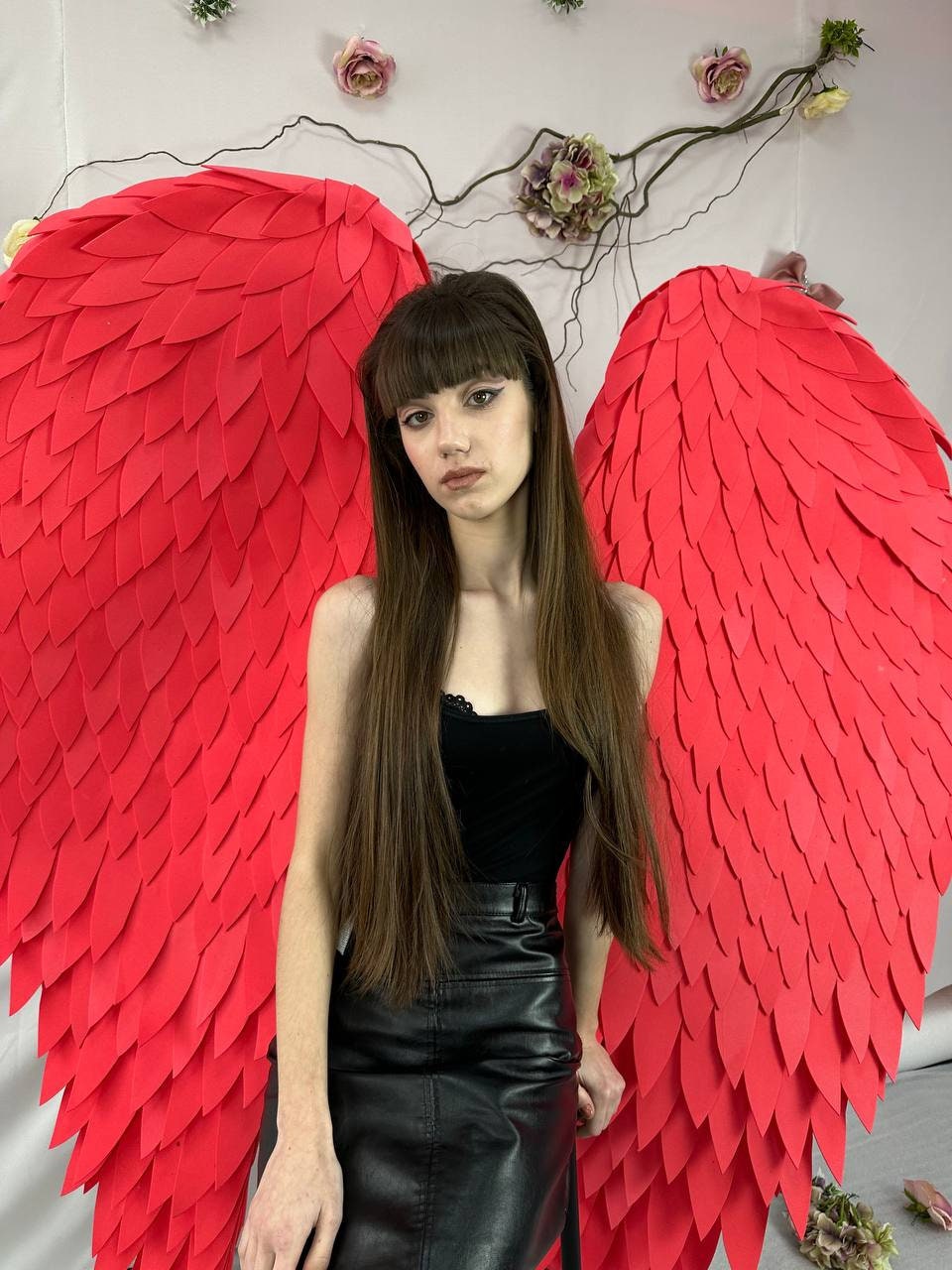 May include: A person wearing a black top and leather skirt, posing in front of large, vibrant red angel wings. The wings are made of layered, textured material. Floral decorations are visible in the background.