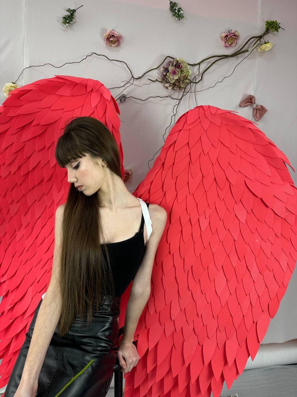 May include: A person wearing a black top and leather skirt with large red angel wings. The wings are made of layered, textured material. The background includes floral decorations.
