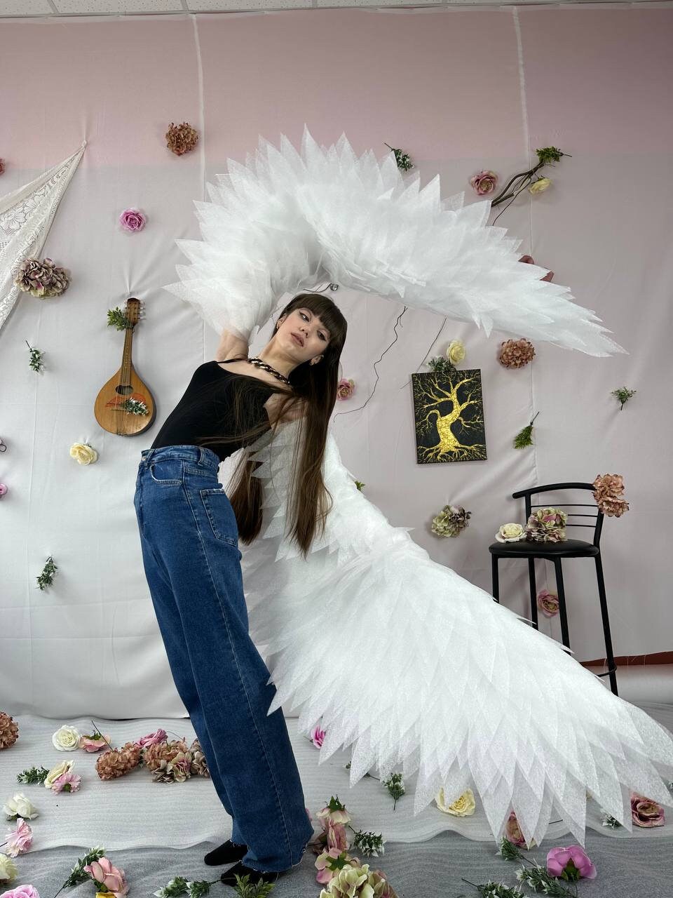 May include: A person wearing a black tank top, blue jeans, and a white feathered angel wing costume. The person is standing in front of a pink wall with white flowers and a guitar.