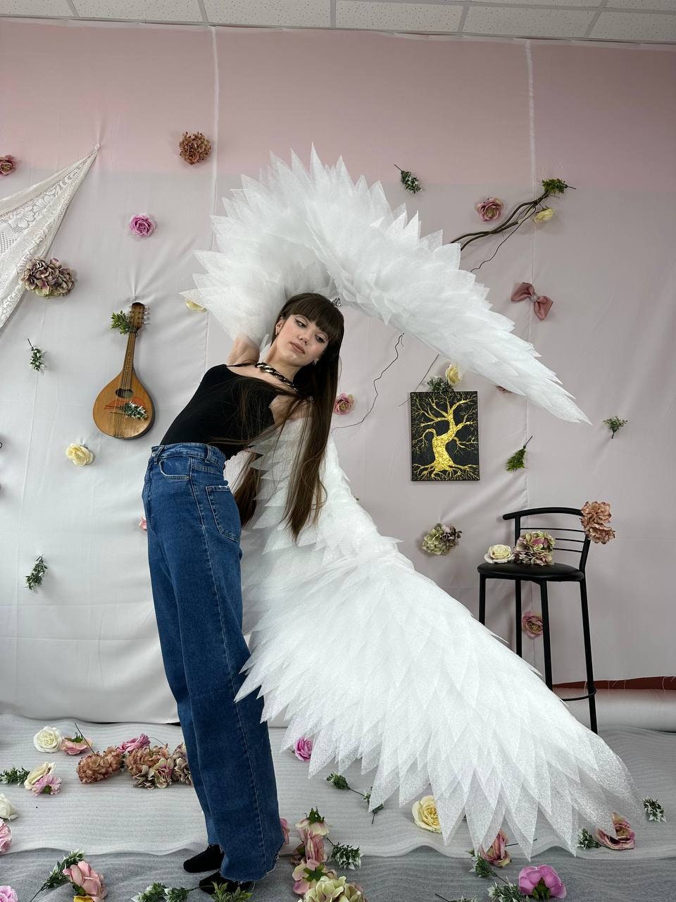 May include: A person wearing a black top and blue jeans with large white feathered wings. The wings are made of many layers of white fabric and are spread out behind the person. The person is standing in front of a pink wall with white flowers and a white lace curtain.