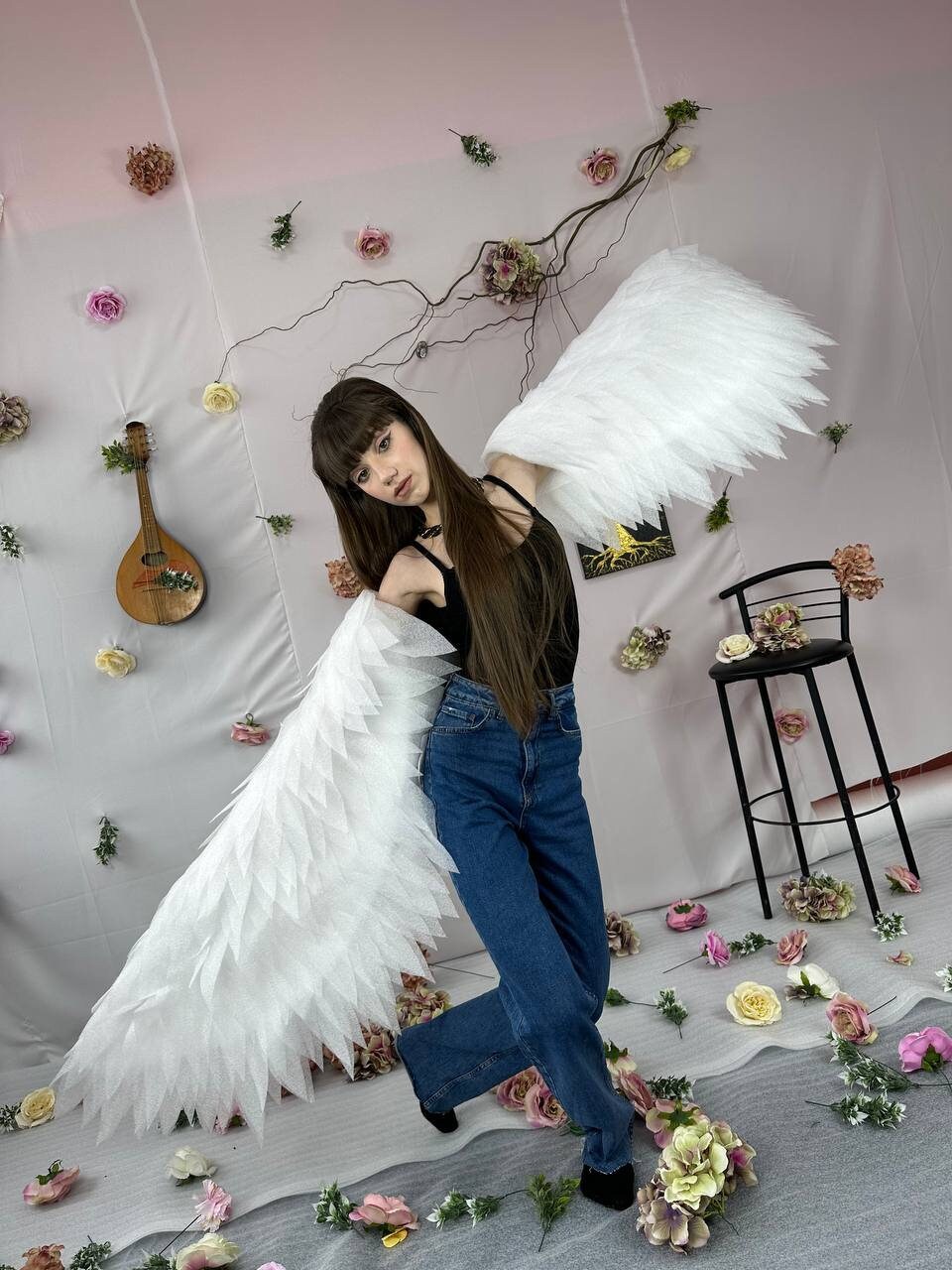 May include: A woman wearing a black tank top and blue jeans poses with large white feathered wings. The background features a pink wall with a vine of artificial flowers.