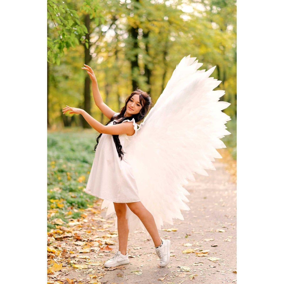 May include: A young woman wearing a white dress and white angel wings poses in a forest setting. The wings are large and feathery, and the woman is smiling. The background is a blur of green and brown leaves.