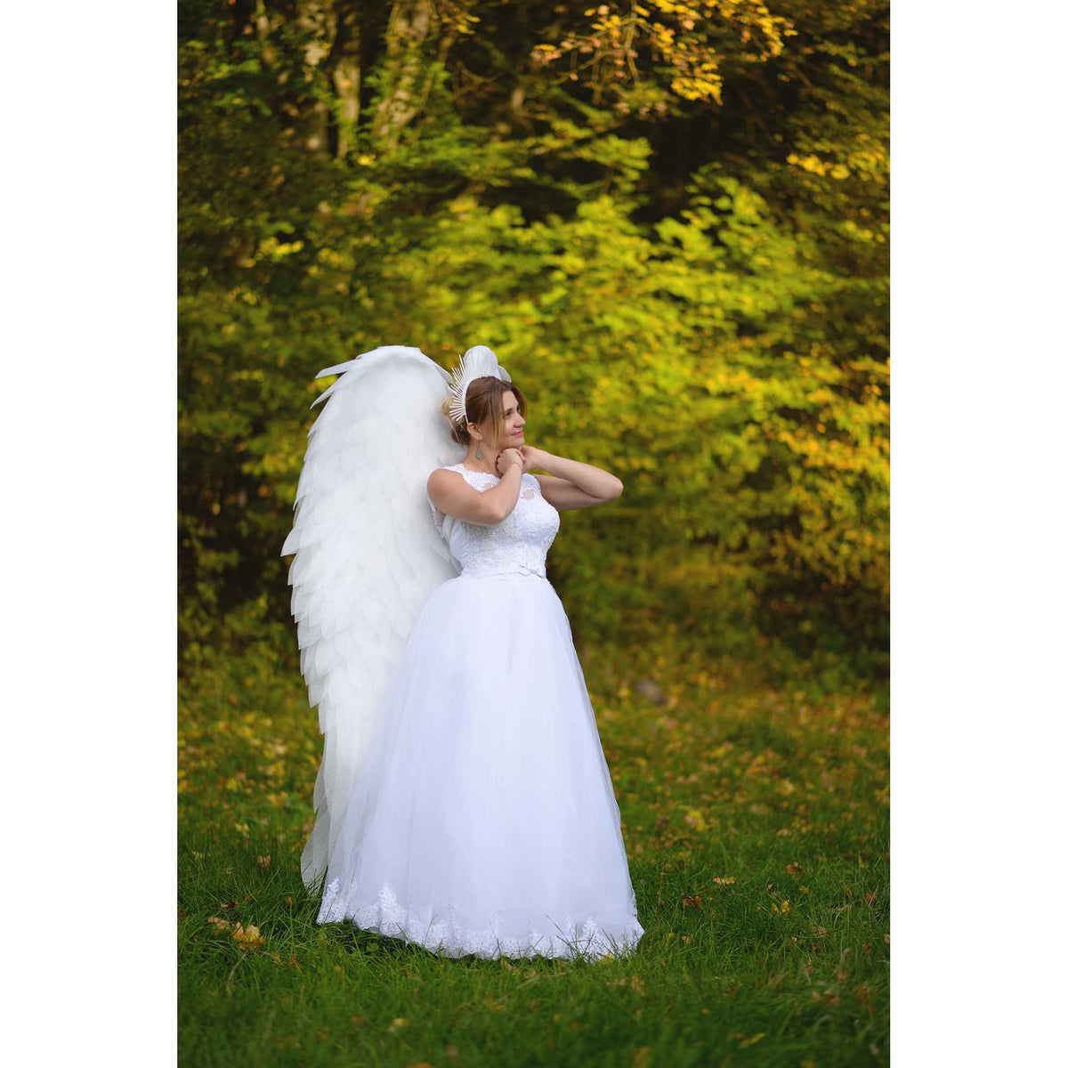 May include: A woman wearing a white dress and large white feathered angel wings stands in a grassy area with a forest background. She is wearing a white dress with lace detailing and has her hands near her face.