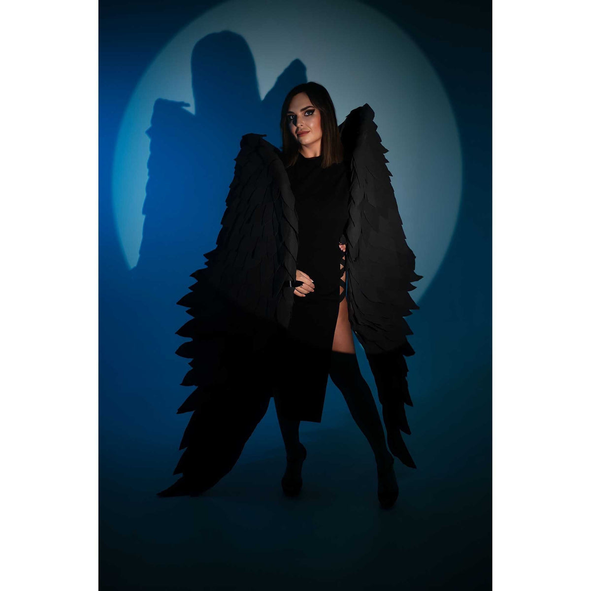 May include: A woman wearing a black dress with a pair of large black feathered wings. The wings are attached to the shoulders and extend behind her. She is standing in a spotlight against a blue background.