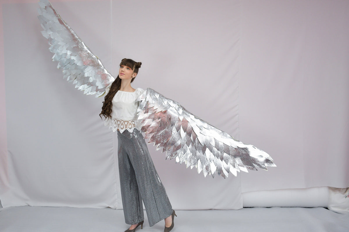 May include: A person wearing a white top and gray pants with large silver wings.
