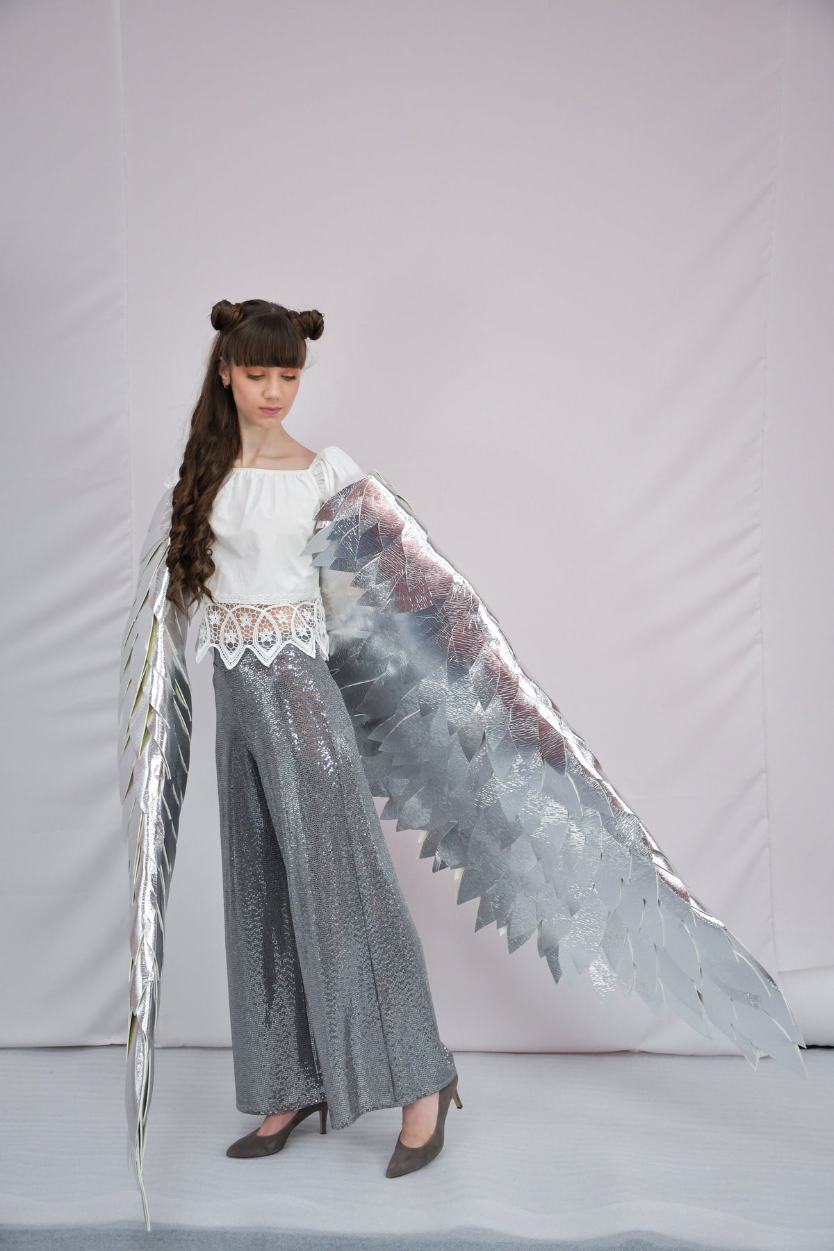 May include: A young person wearing a white top and silver sequined pants with large silver wings. The wings are made of many individual pieces of silver material.