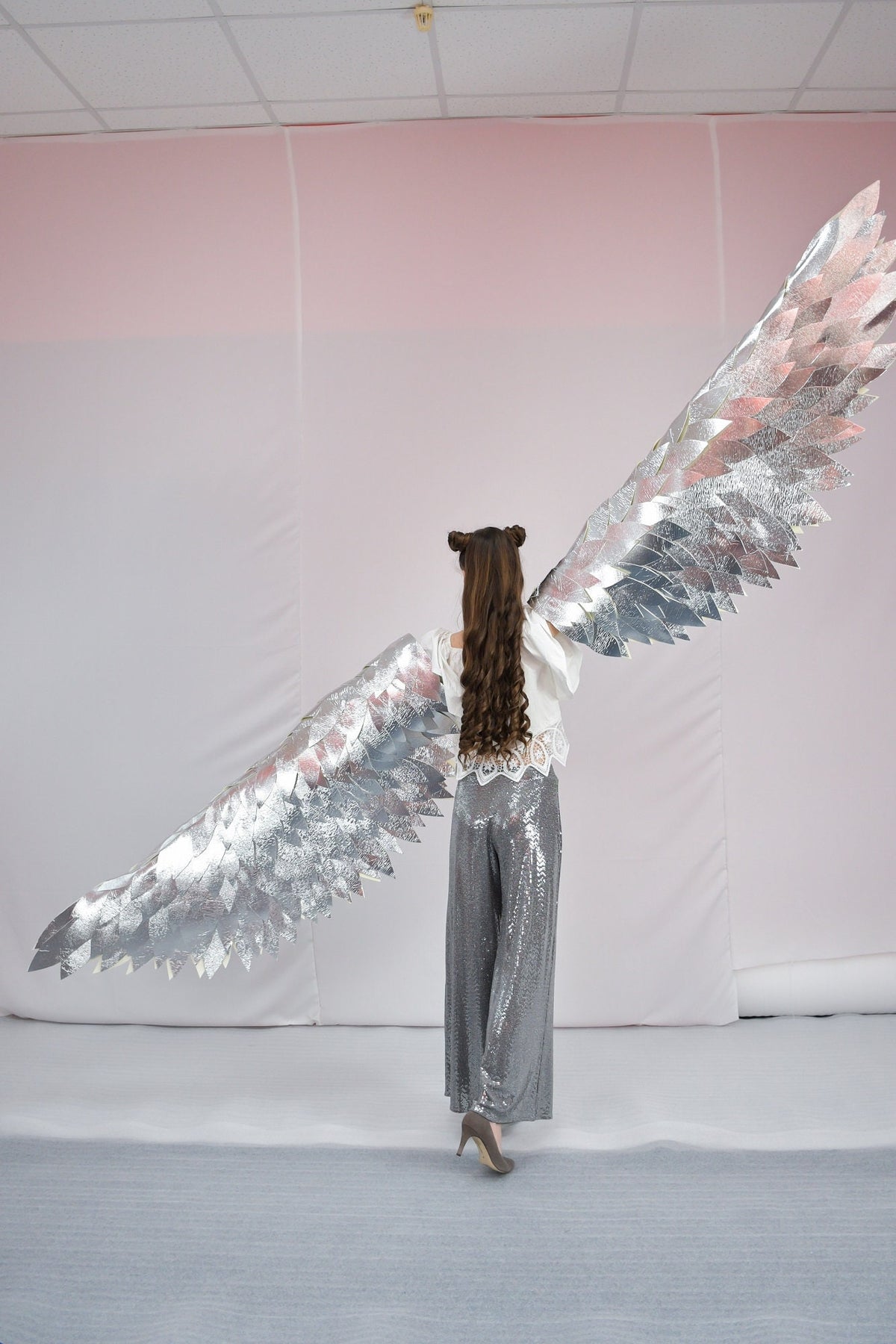 May include: A person wearing a white top and silver sequined pants with large silver wings behind them. The wings are made of many small, overlapping pieces of silver material.