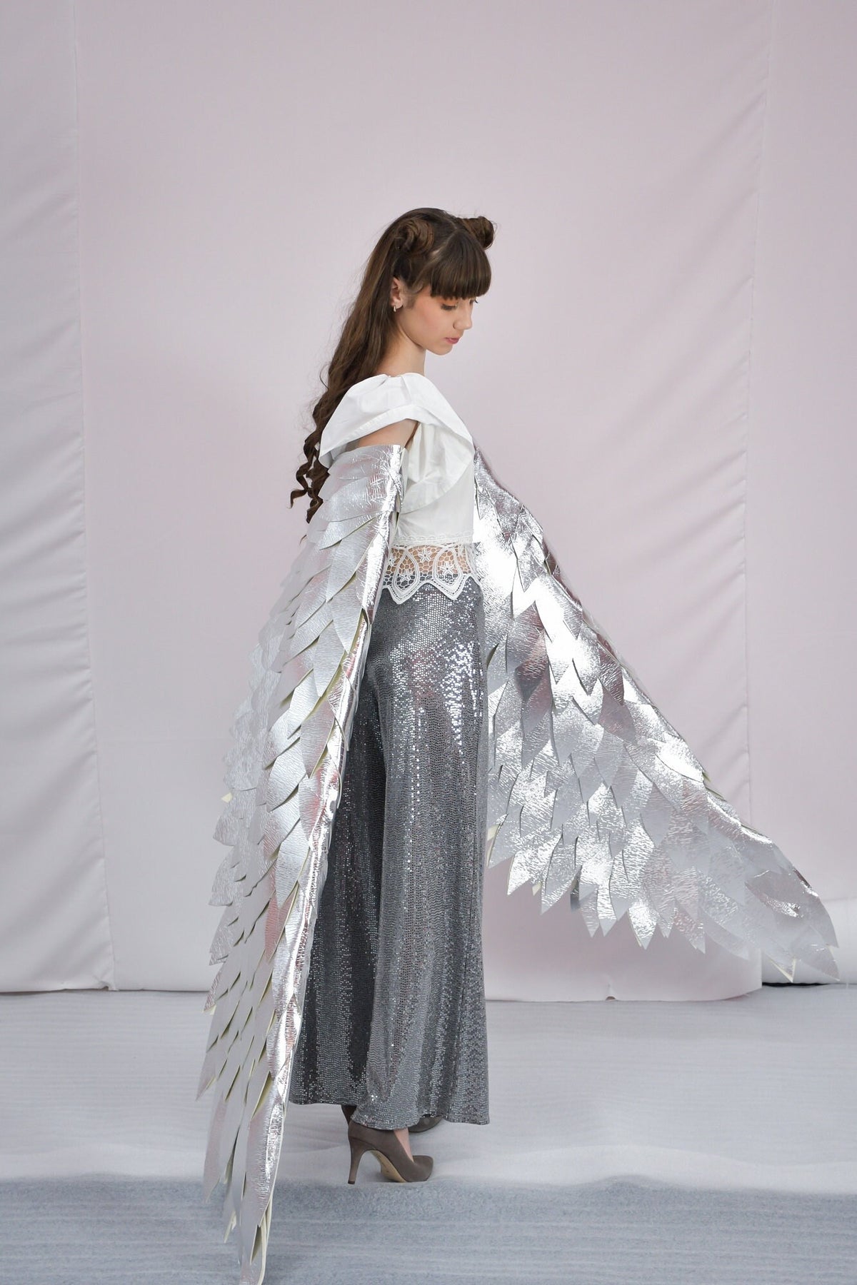 May include: A woman wearing a white top with silver sequined pants and large silver wings. The wings are made of feathers and are attached to the woman's shoulders.