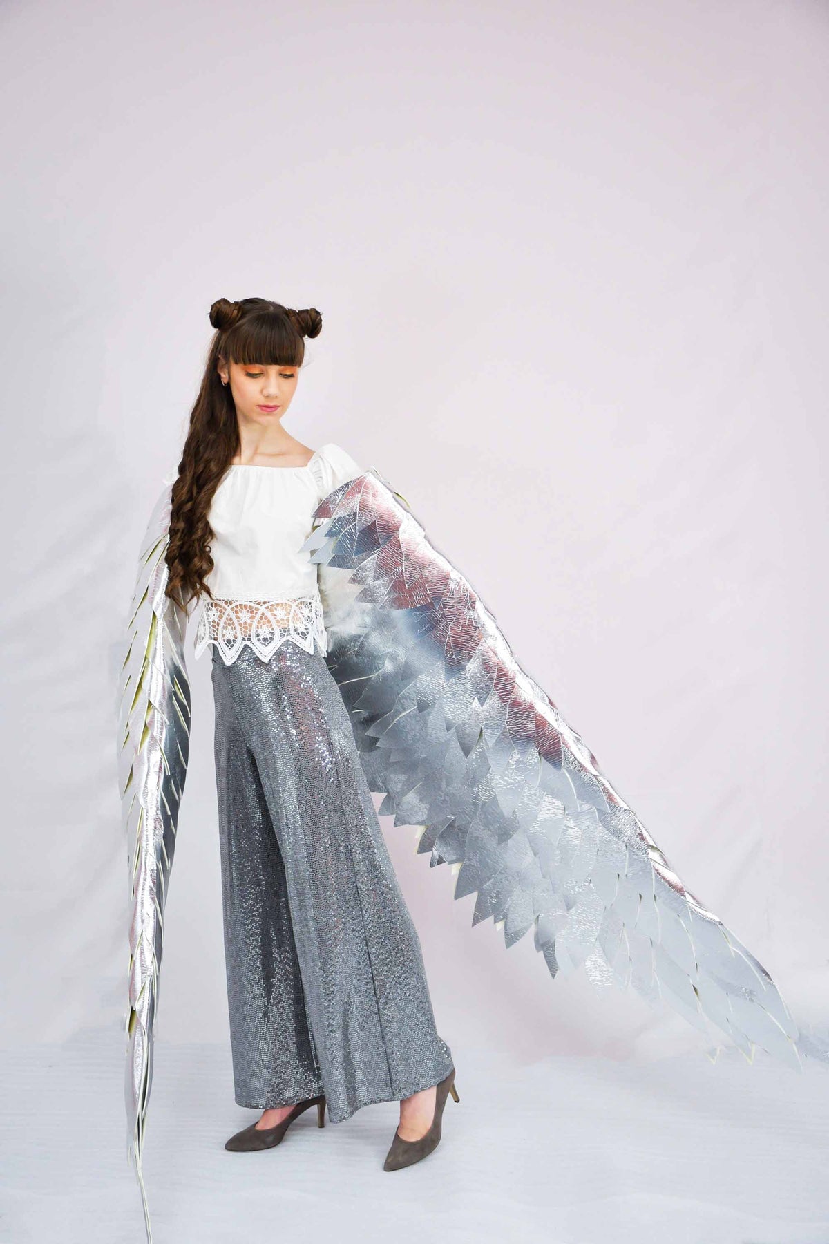 May include: A young person wearing a white top and gray sequined pants with large silver wings.