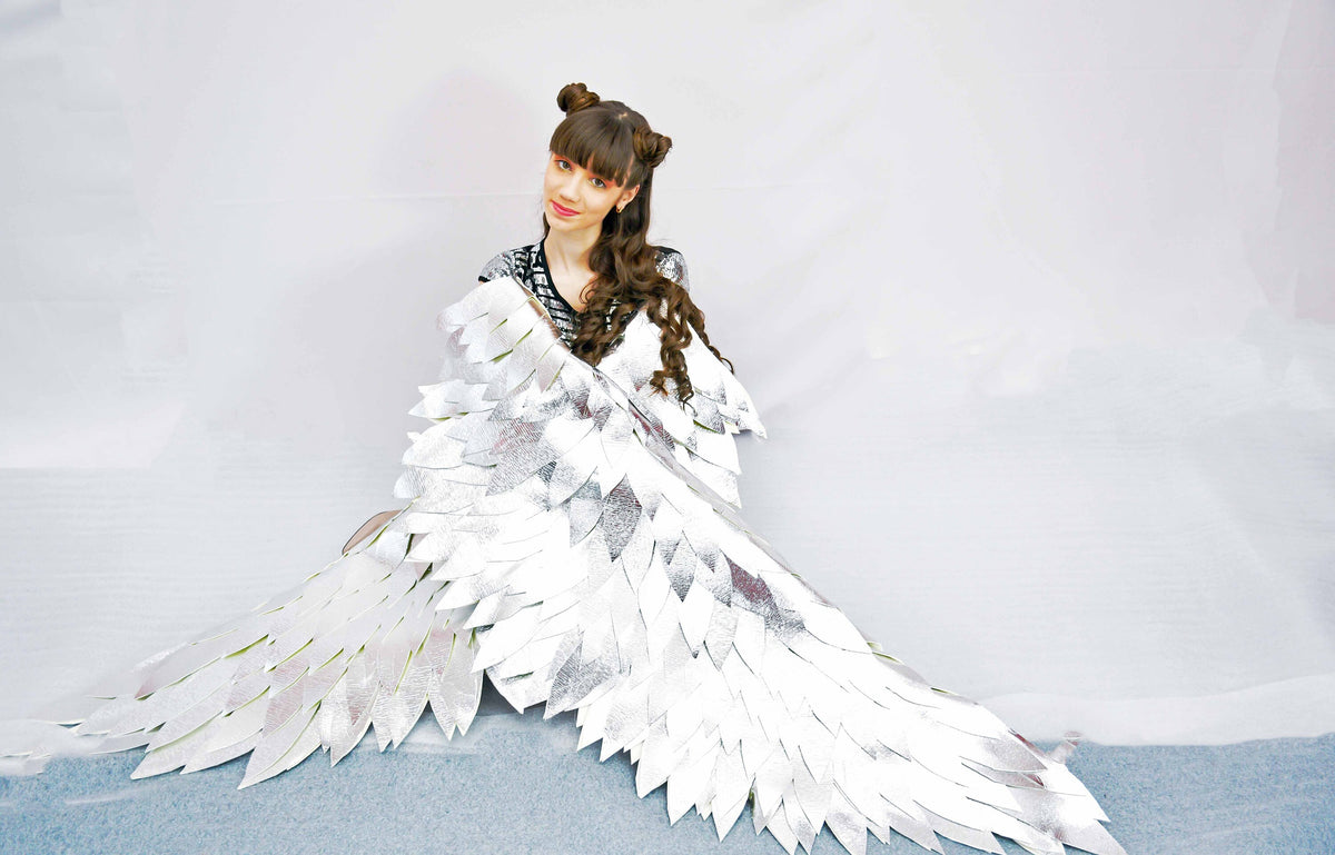 May include: A woman wearing a silver costume with large, feathery wings. The wings are made of many individual feathers that are arranged in a layered pattern. The woman is sitting on a white surface and looking at the camera.