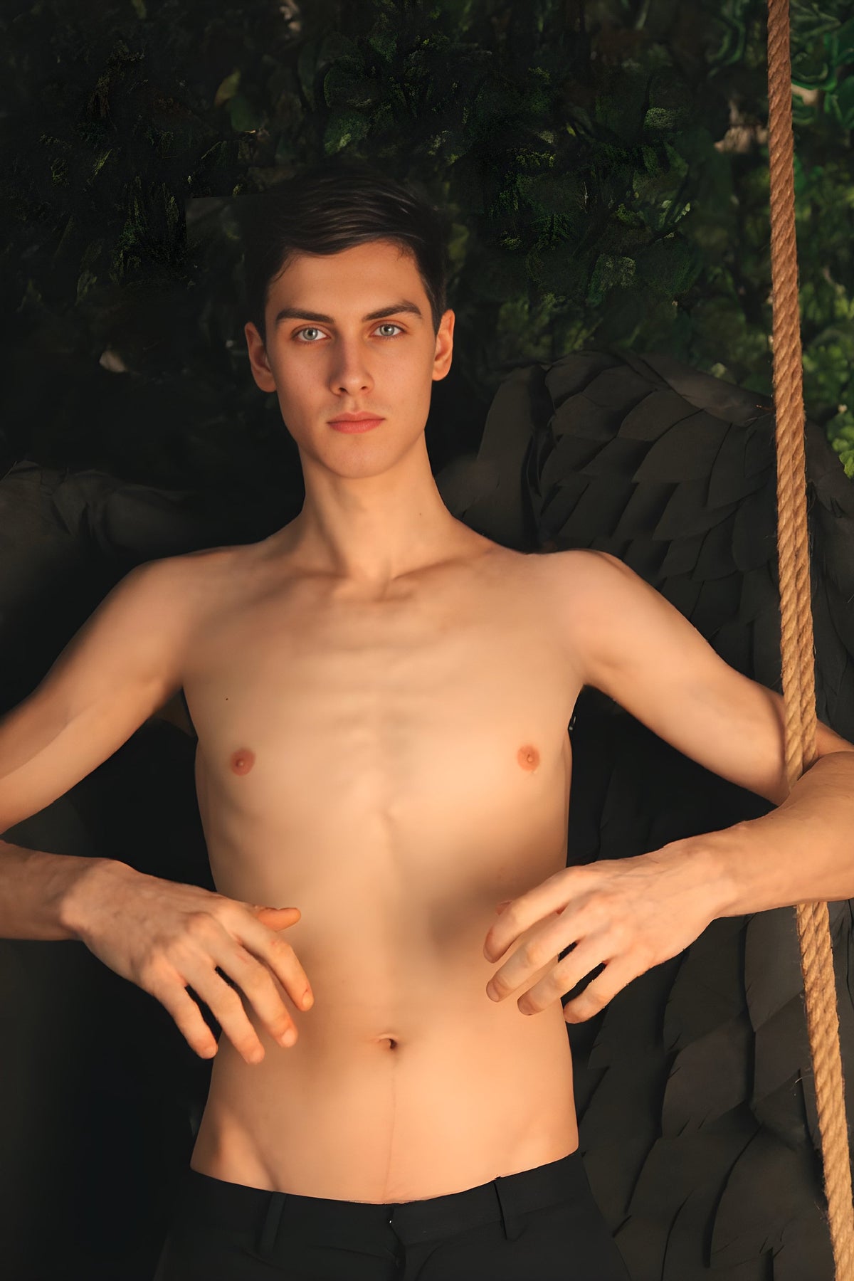 May include: A shirtless man with short brown hair and blue eyes sits with his arms crossed in front of him. He is wearing black pants and is sitting in front of a black background.