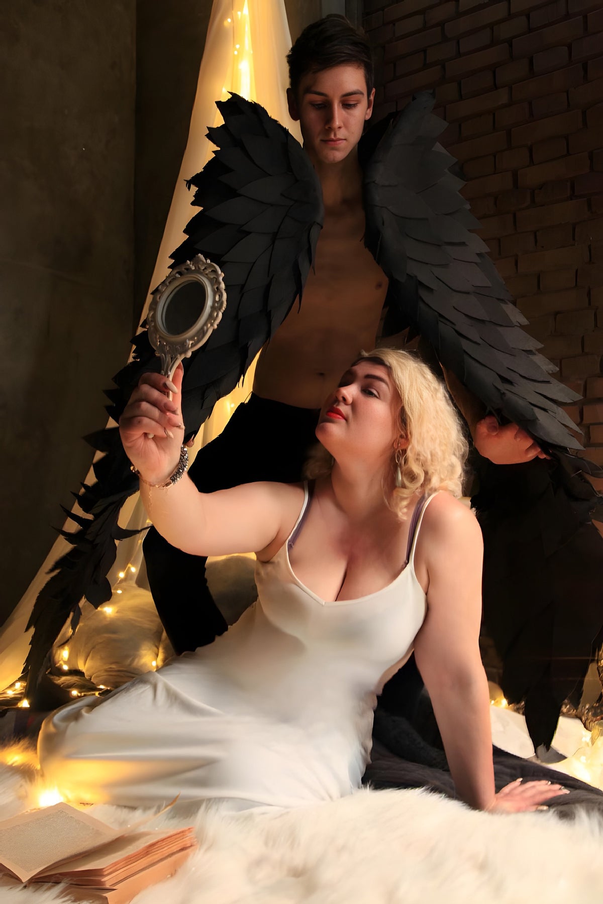 May include: A woman in a white dress holds a hand mirror and looks at her reflection. A man with black wings stands behind her. The scene is lit with warm, golden lights.