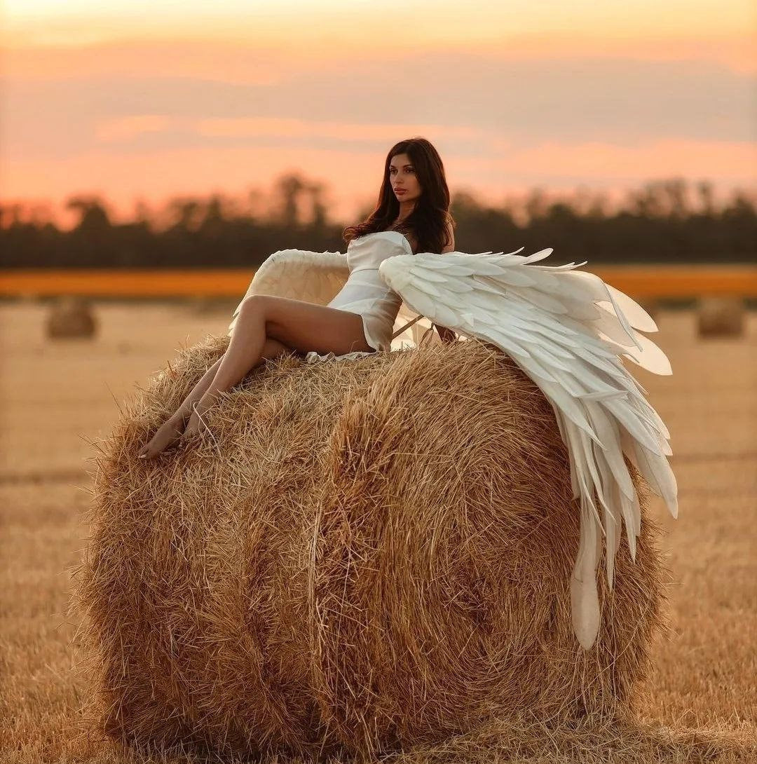May include: A woman wearing a white dress sits on a large bale of hay with a pair of large white angel wings draped over her shoulders.