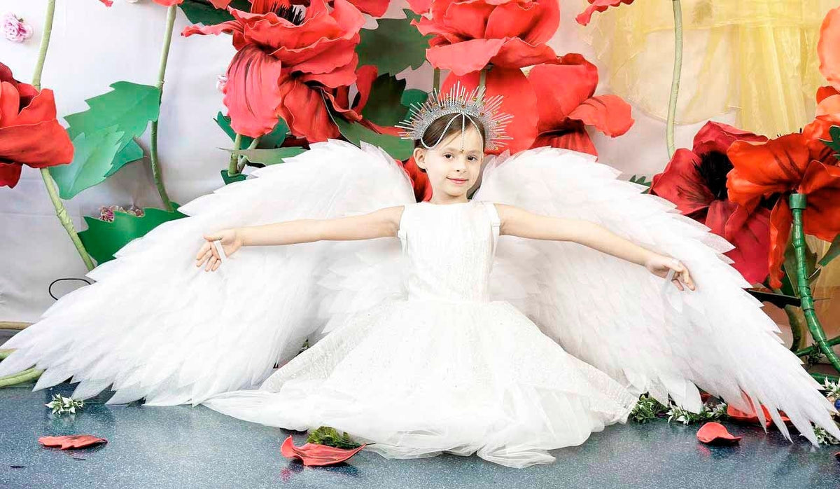 May include: A young person dressed as an angel, wearing a white dress and large feathered wings. They are posed with arms outstretched, in front of large red paper flowers. A silver halo-like headpiece completes the angelic look.