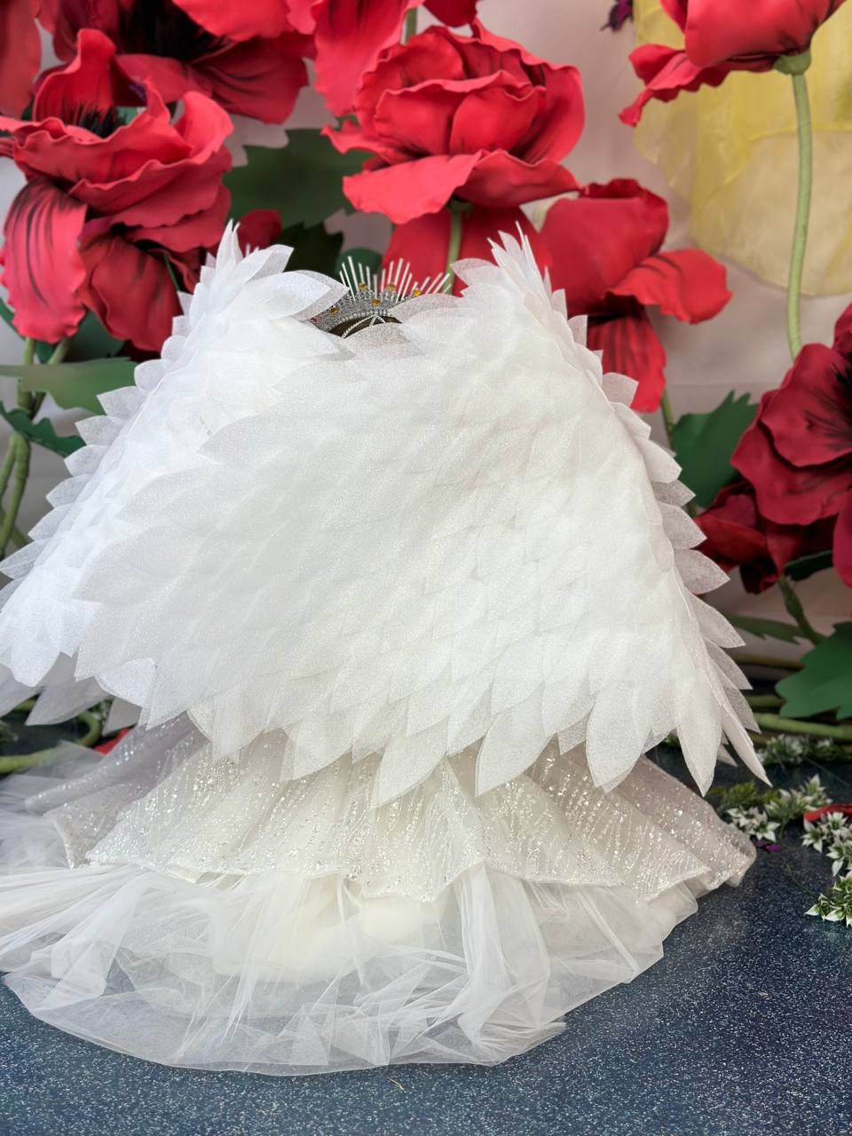 May include: A white, layered costume with a feathery wing design. The costume has a tiered skirt and a small, jeweled crown. The background features large red flowers.