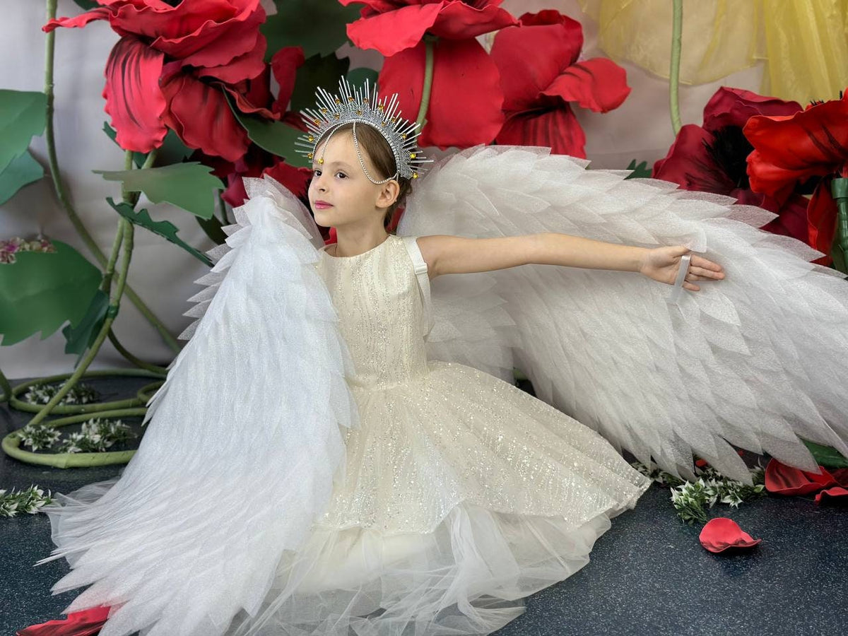 May include: A young person wearing a cream-colored dress with a layered skirt and a silver crown. Large white feathered wings are attached to the back. The background features large red flower decorations and green foliage.