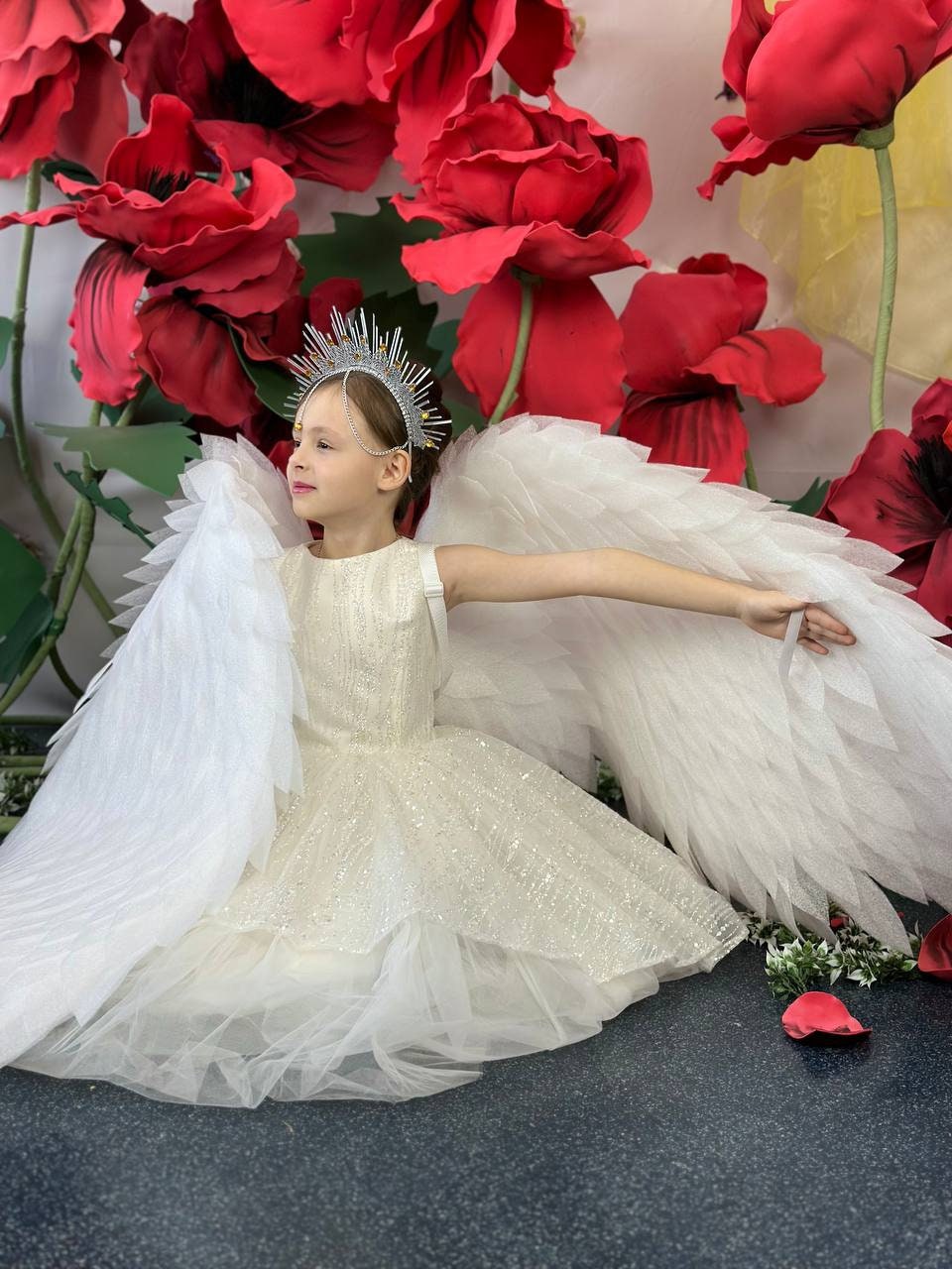 May include: A young person wearing a cream-colored, sparkly dress with white feathered wings and a silver crown. The background features large red and yellow paper flowers, creating a whimsical and angelic theme.