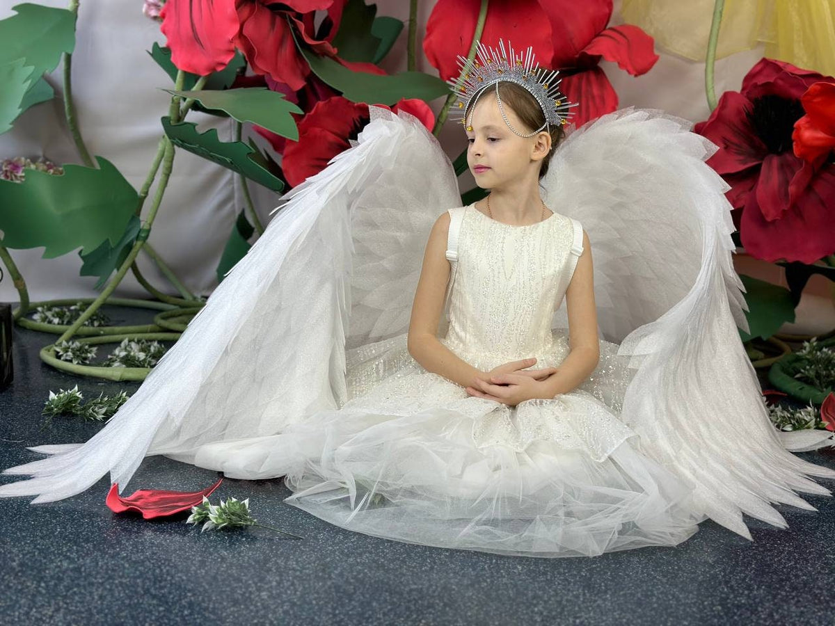 May include: A young person wearing a white dress, silver headpiece, and large white angel wings. The wings are spread out behind the person, and the dress has a layered skirt. The background includes large red flowers and green foliage.