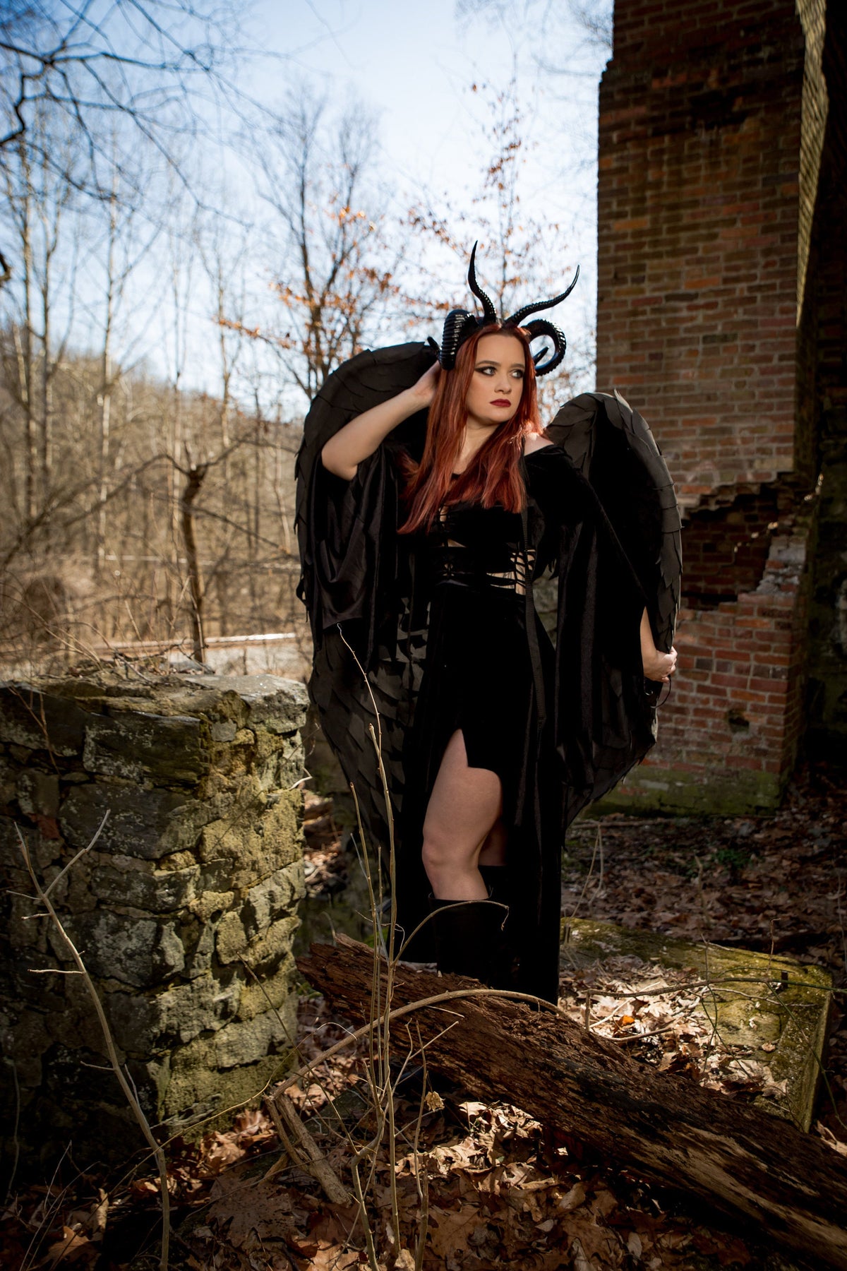 May include: A woman wearing a black dress with a black feathered wings and horns. She is standing in a forest setting with a stone wall behind her.