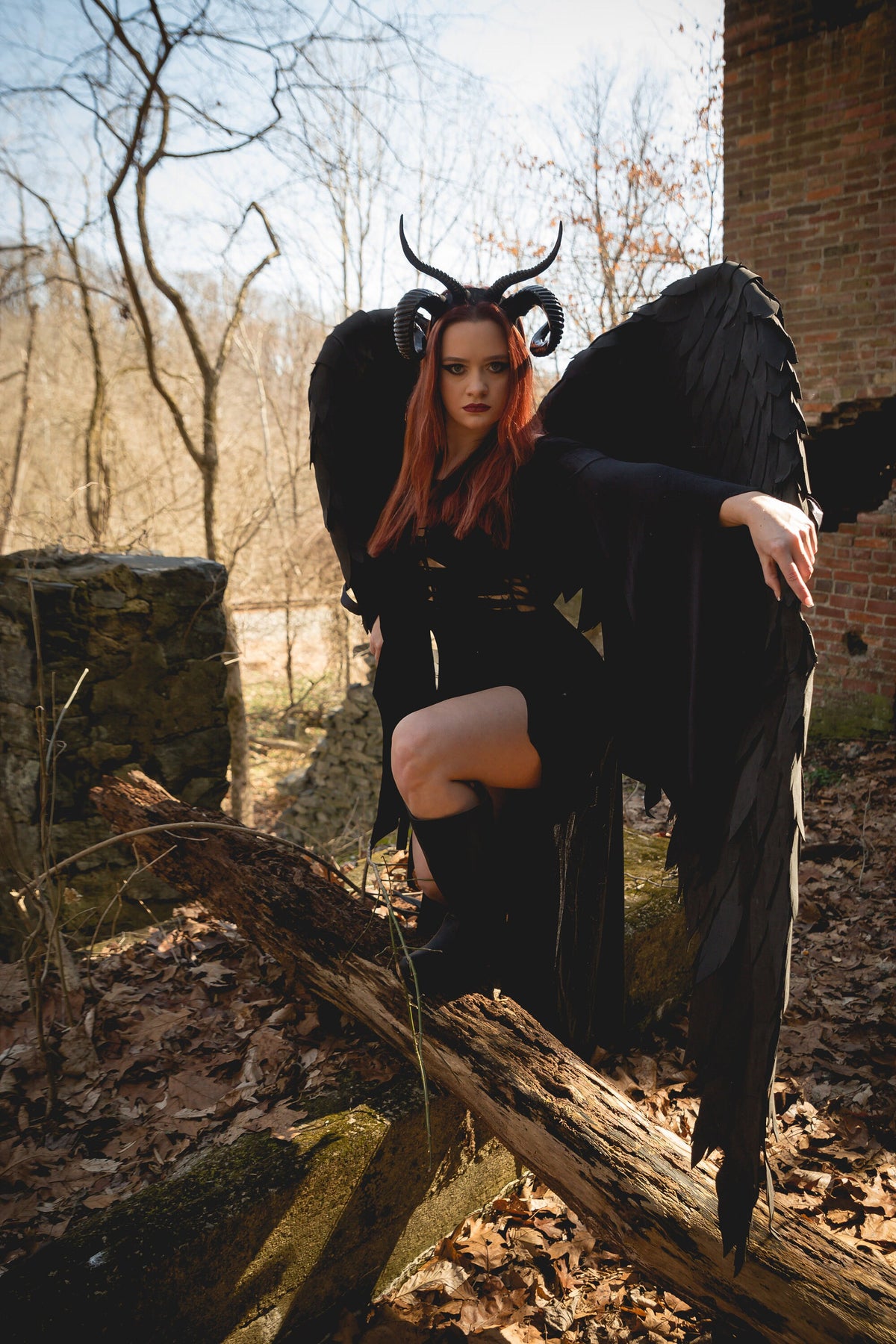May include: A woman with red hair wearing a black dress and large black wings with horns on her head. She is standing in a forest setting with a stone wall and a fallen log in the background.