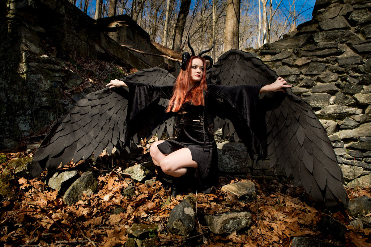 May include: A woman with red hair and horns is wearing a black dress and large black wings. She is sitting on a pile of rocks in a forest setting.