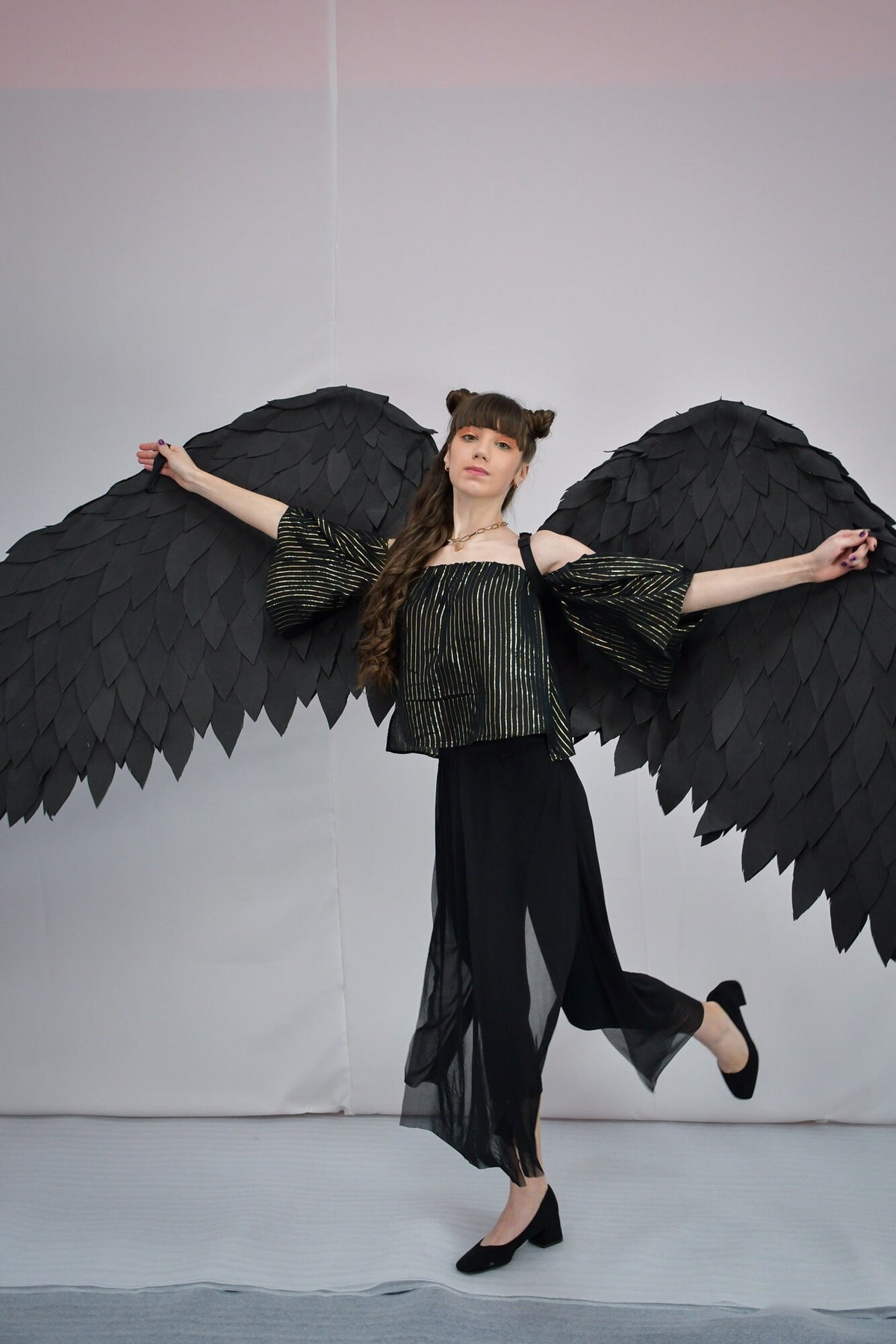 May include: A woman wearing a black and gold striped top and black pants with a sheer overlay, poses with large black angel wings.