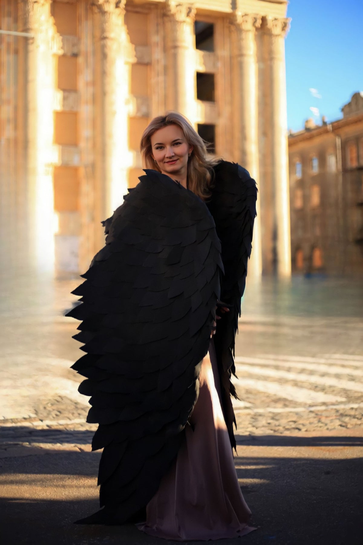 May include: A woman wearing a long, flowing dress with large black feathered wings. The wings are made of many individual feathers and are very detailed. The woman is standing in front of a building with columns.