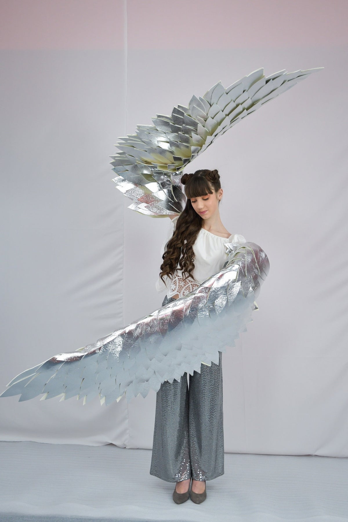 May include: A person wearing a white top and silver sequined pants is holding large silver wings made of paper. The wings are shaped like a bird's wings and are spread out behind the person.