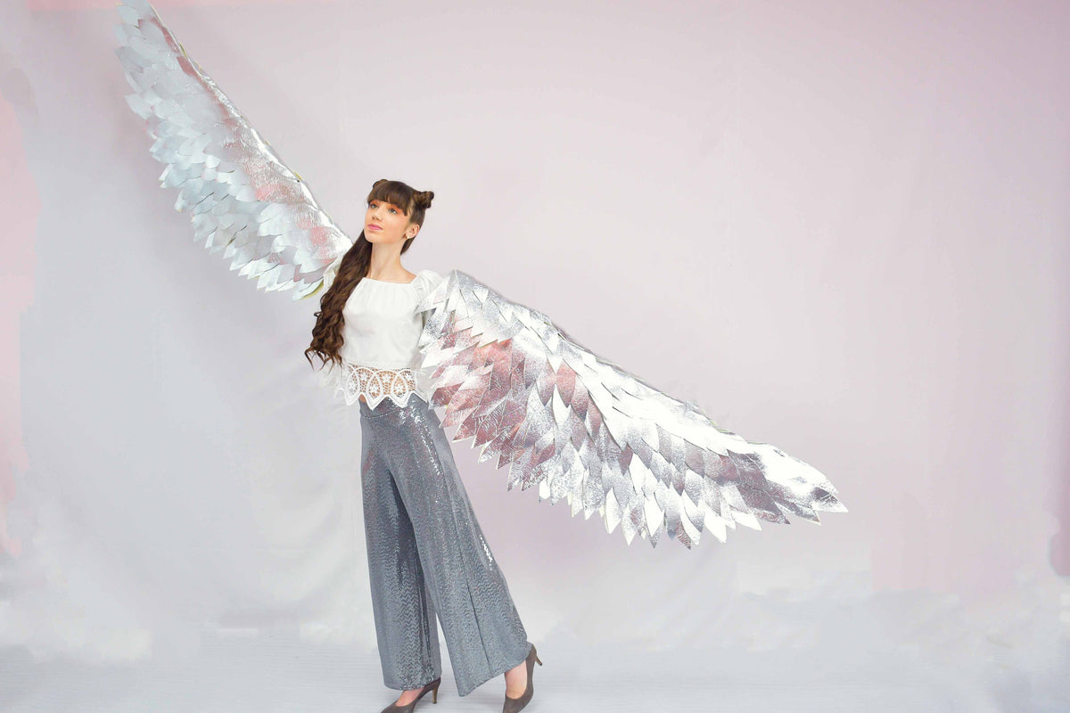 May include: A woman wearing a white top and gray sequined pants poses with large silver wings. The wings are made of feathers and are spread out behind her.