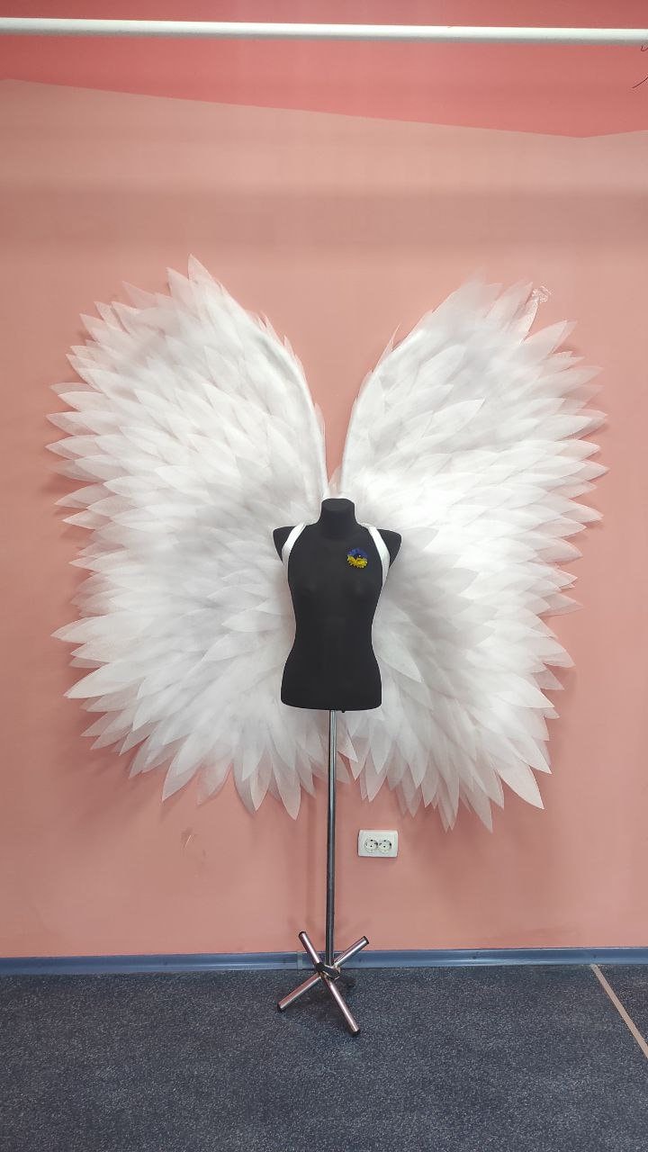 May include: A black mannequin with white straps and a yellow and blue flower pin stands in front of large white angel wings. The wings are made of layered, feathery material. The background is a light pink wall.