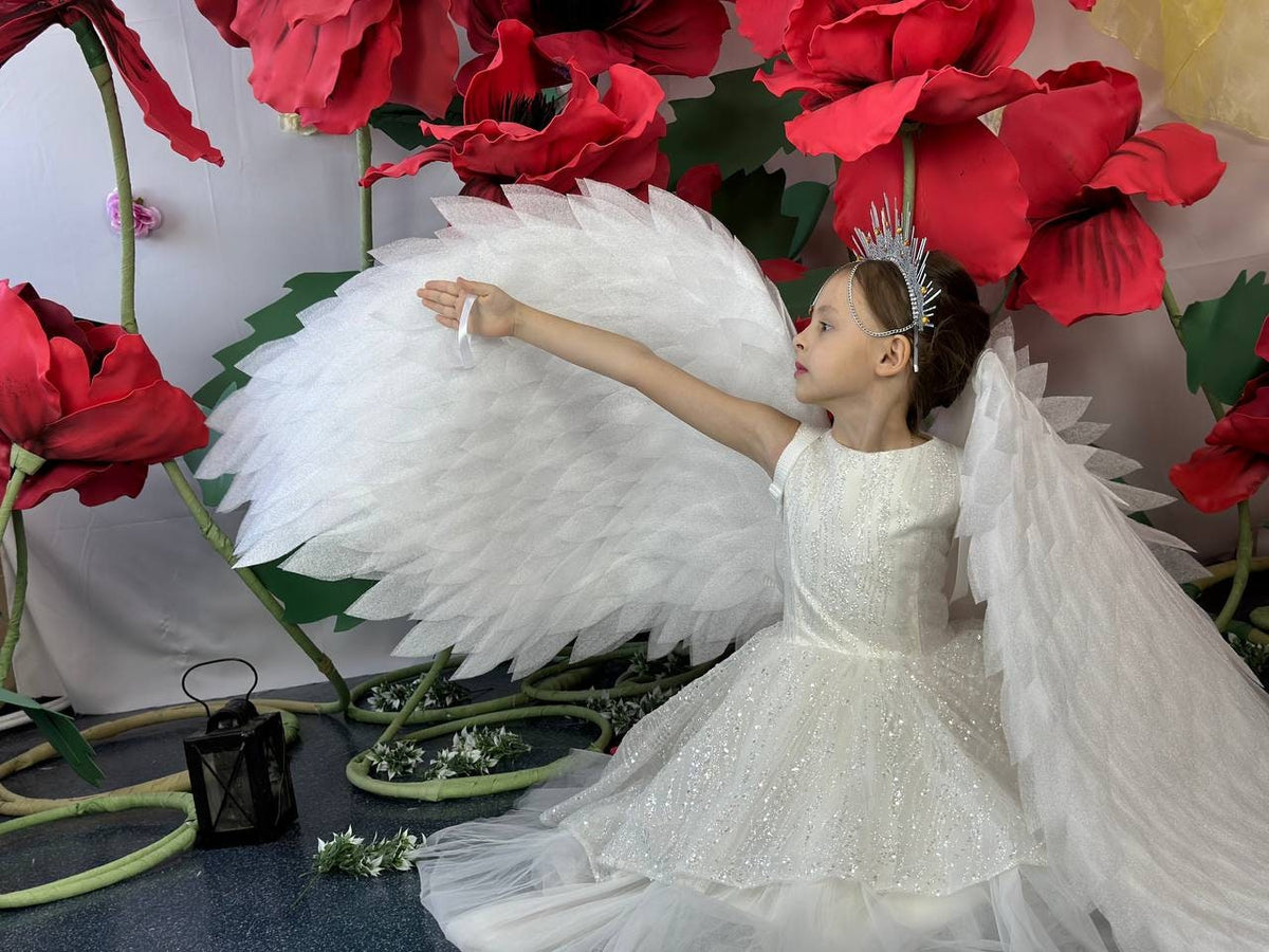May include: A young person in a white dress with a sparkling bodice and a layered skirt, wearing a silver tiara and large white feathered wings. The background features oversized red flowers and green foliage.