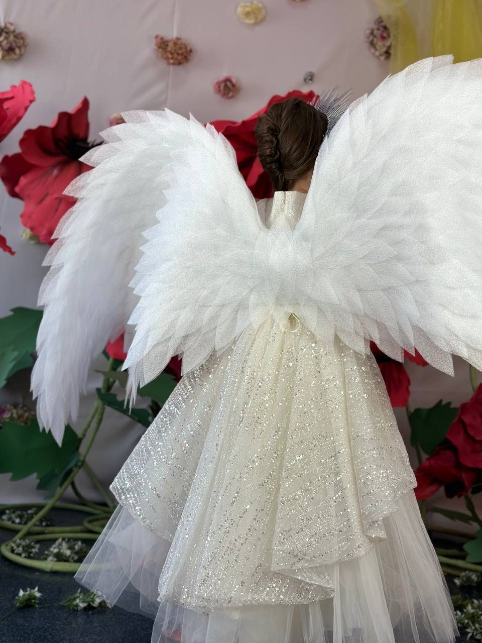 May include: A white angel costume featuring large, layered wings and a shimmering, layered dress. The wings are made of multiple layers of white fabric. The dress has a sparkly overlay. The background includes large red flower decorations.