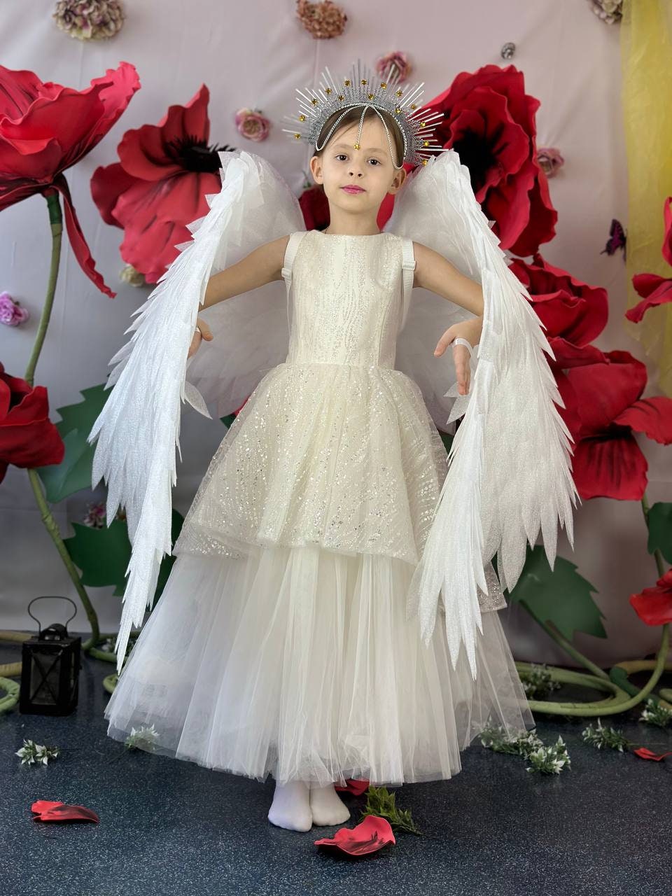 May include: A young person in a white angel costume with large feathered wings and a silver halo. The dress is layered with a sparkly bodice and a flowing tulle skirt. The background features large red flower decorations.