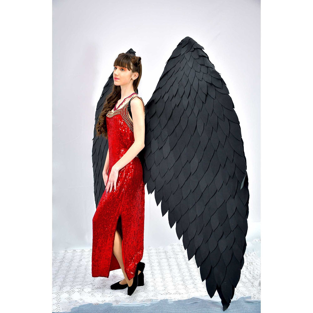 May include: A woman wearing a red sequined dress with a black feather angel wing costume accessory. The wings are large and dramatic, with a black feather design.
