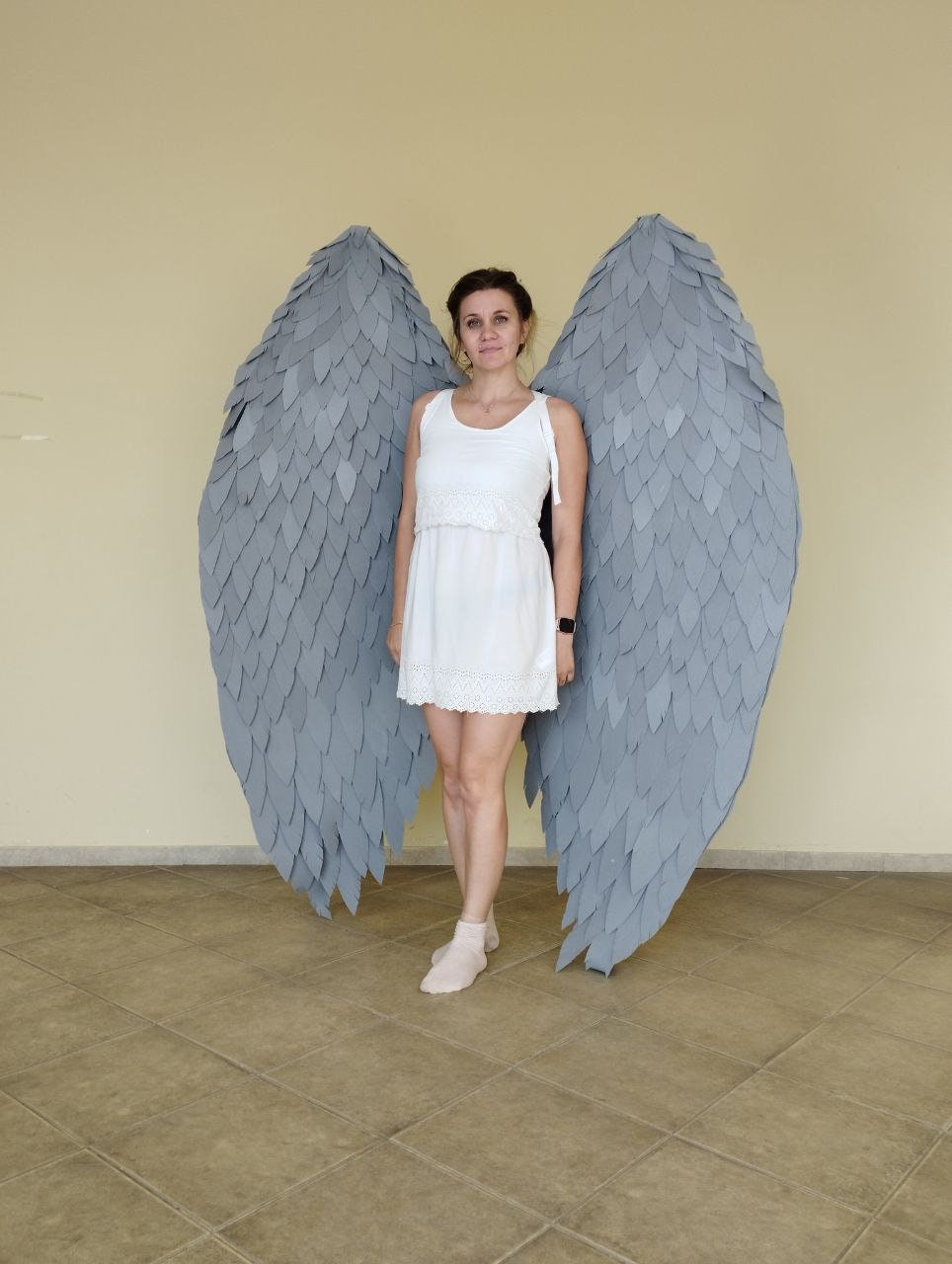 May include: A person wearing a white dress stands in front of a pair of large, gray angel wings. The wings are made of feathers and are attached to a harness.
