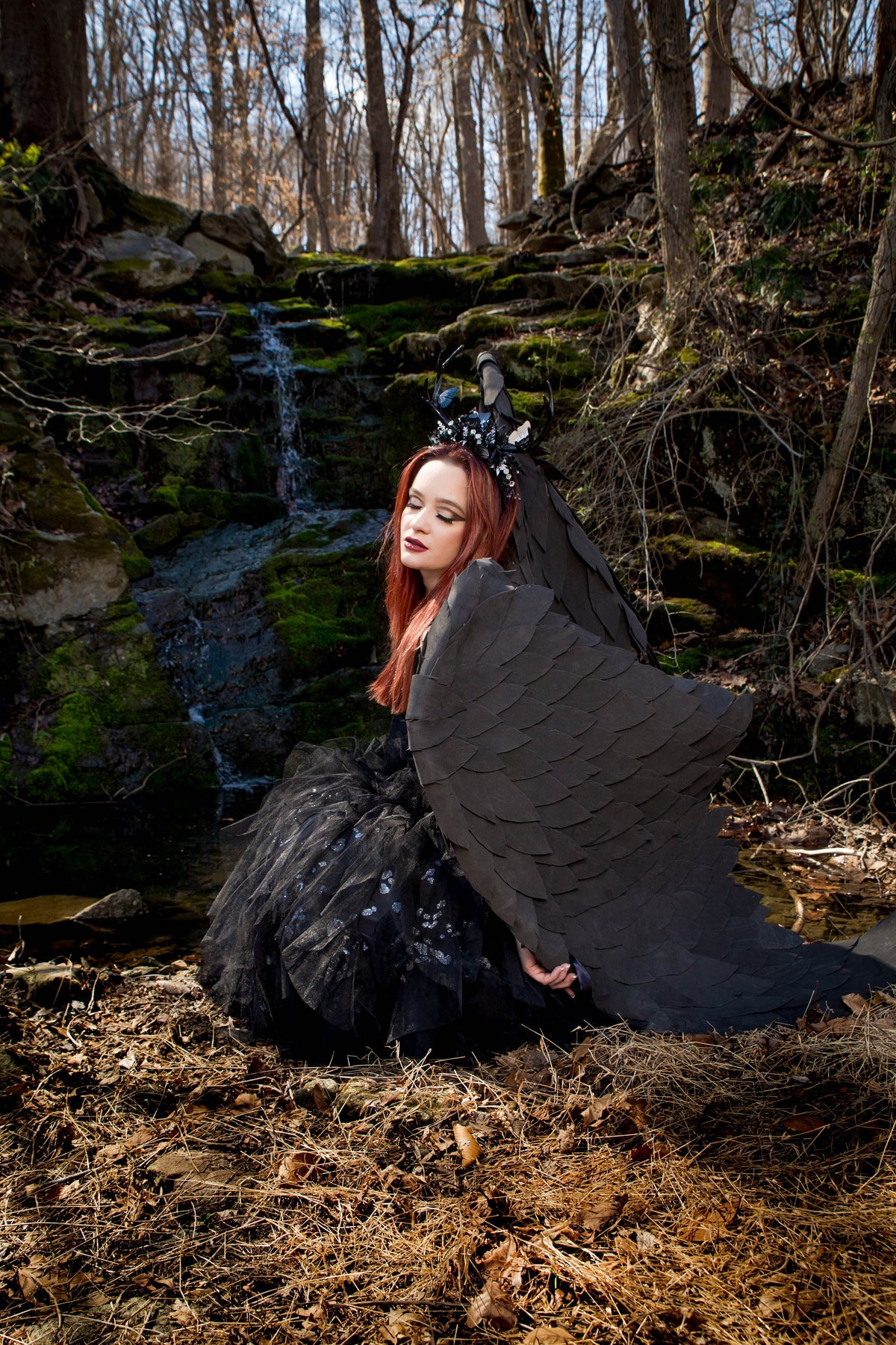 May include: A woman with red hair wearing a black dress and large black wings sits by a small waterfall in a forest setting.
