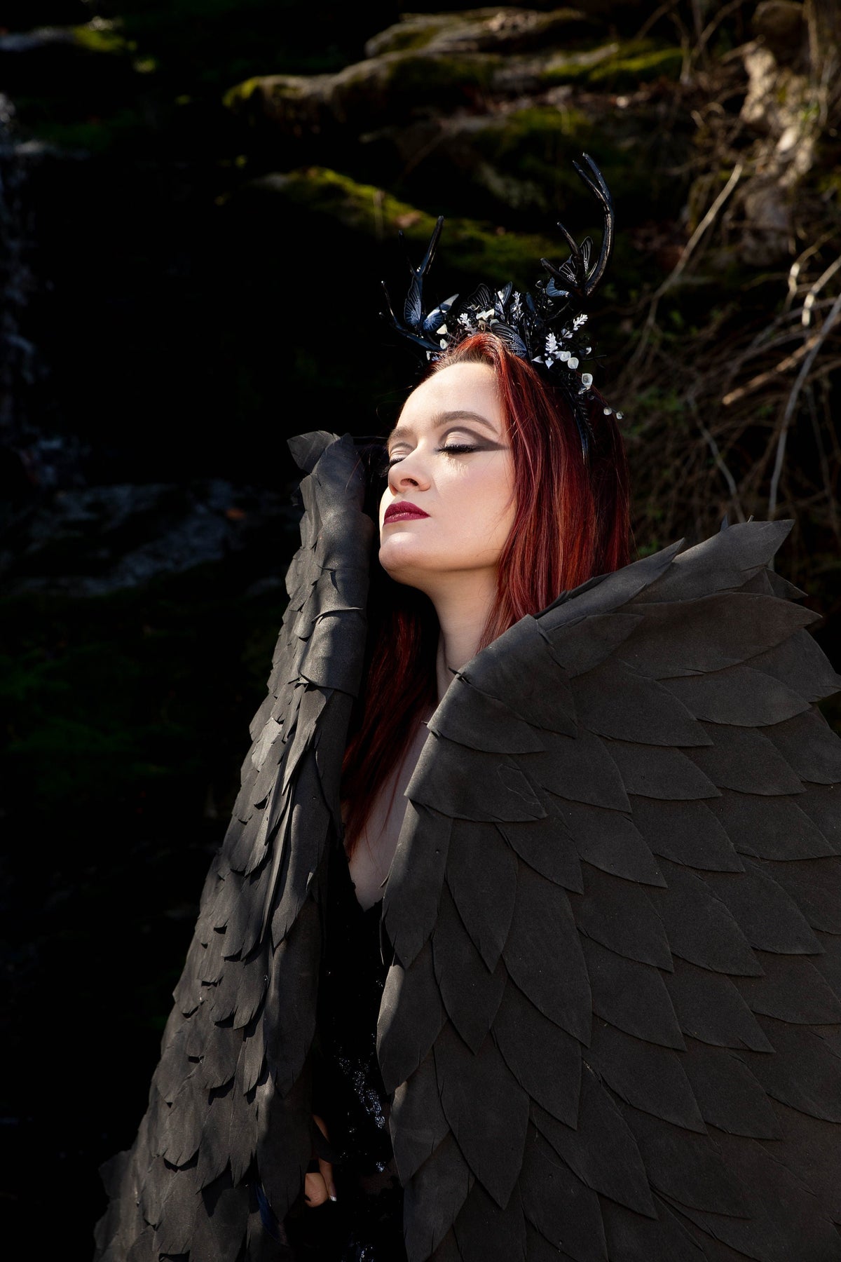 May include: A woman with red hair wearing a black feathered costume with a crown of antlers and rhinestones. The costume has large black wings.