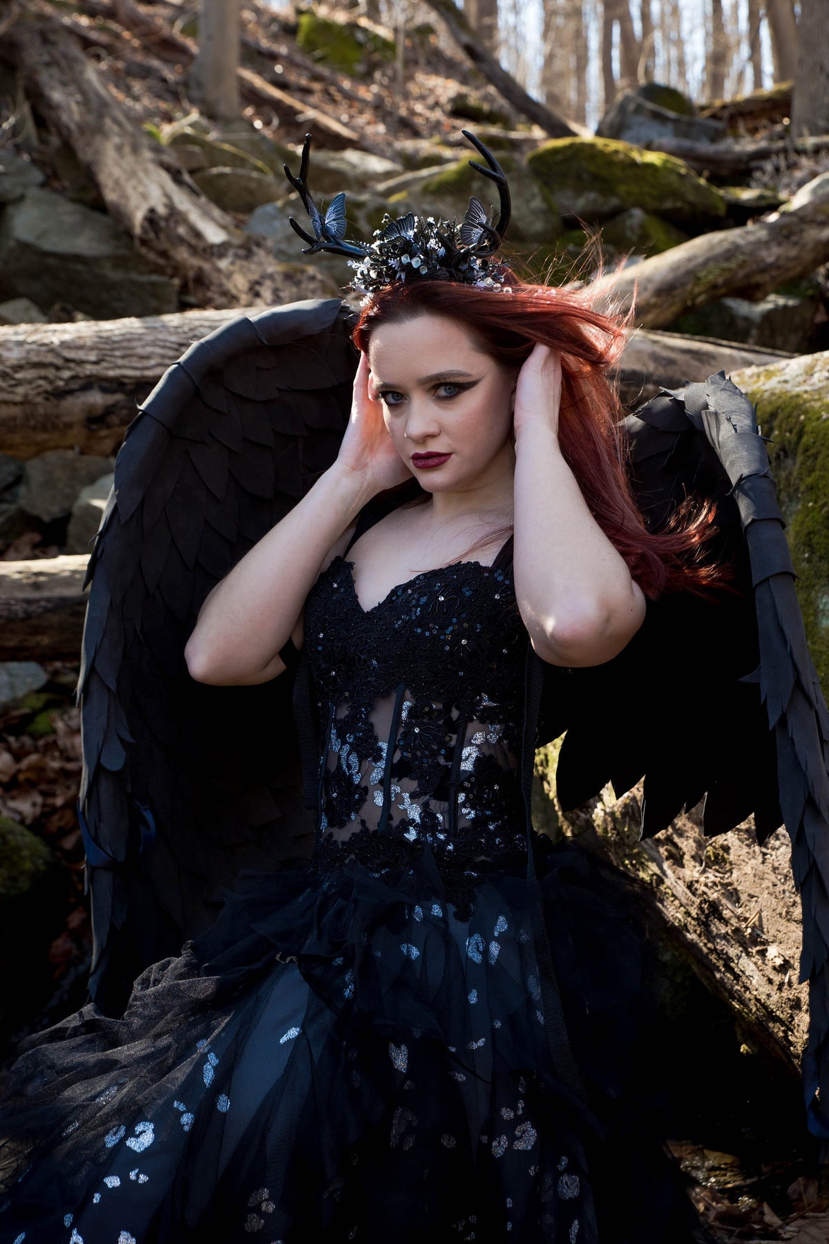 May include: A woman wearing a black dress with a lace bodice and a crown of antlers and butterflies. She is wearing black wings and has red hair. The background is a forest setting.