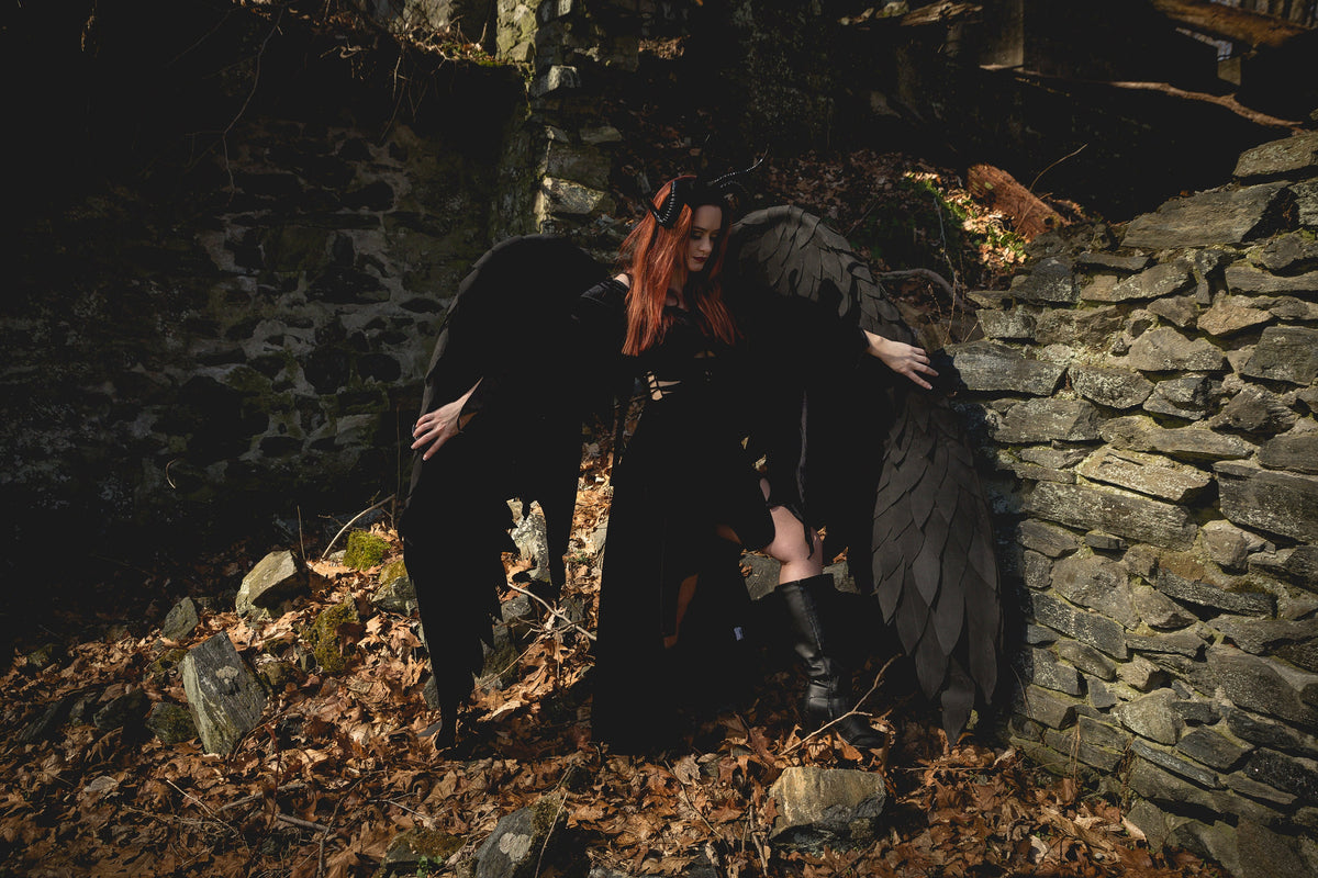 May include: A woman with red hair wearing a black dress and large black wings stands in front of a stone wall. The woman is looking down at the ground. The background is dark and shadowy.