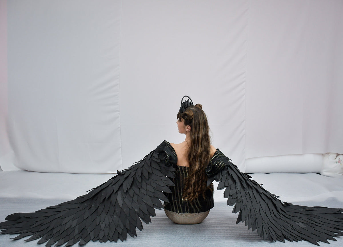 May include: A person wearing a black and gold outfit with large black wings made of feathers. The person is sitting on a white surface with a white background.