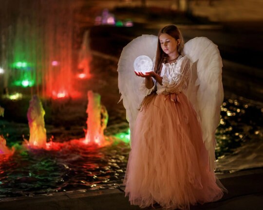May include: A young person wearing a white dress and a white angel wing costume holds a glowing orb in front of a fountain with colorful lights.
